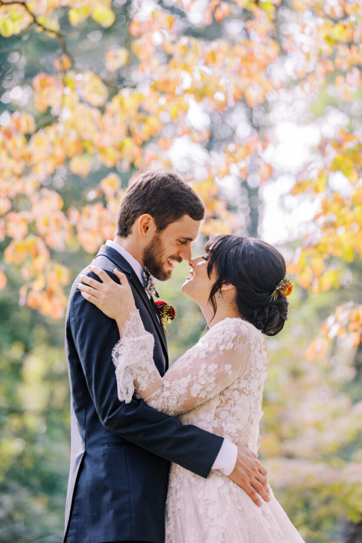 A bride and groom, dressed in formal wedding attire, embrace outdoors with autumn foliage in the background.