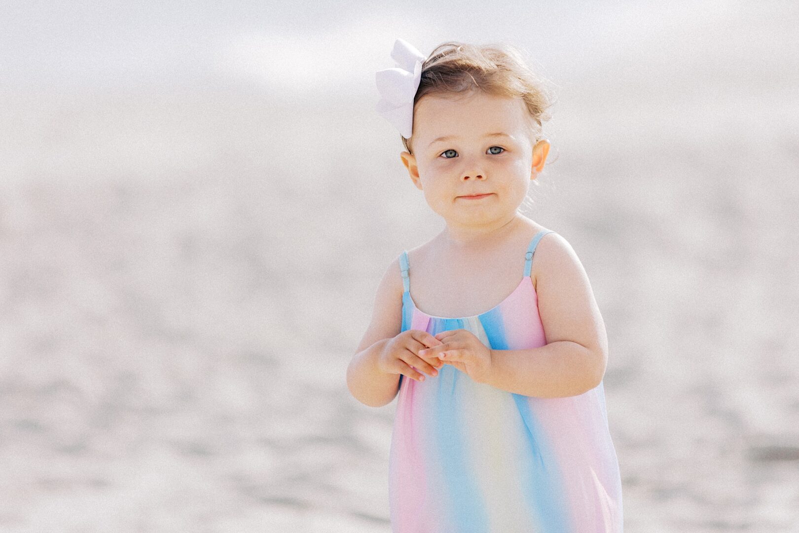 A young child wearing a pastel rainbow dress and a white bow stands on a sandy beach, looking at the camera.