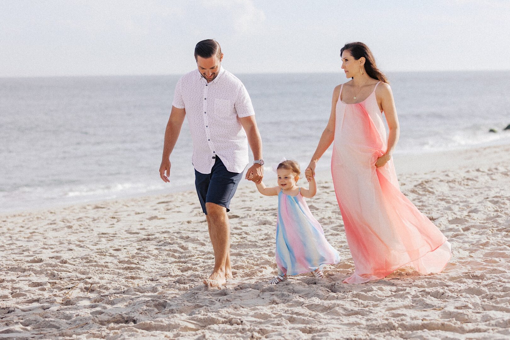 A man, woman, and young girl walk barefoot together on a sandy beach near the ocean.