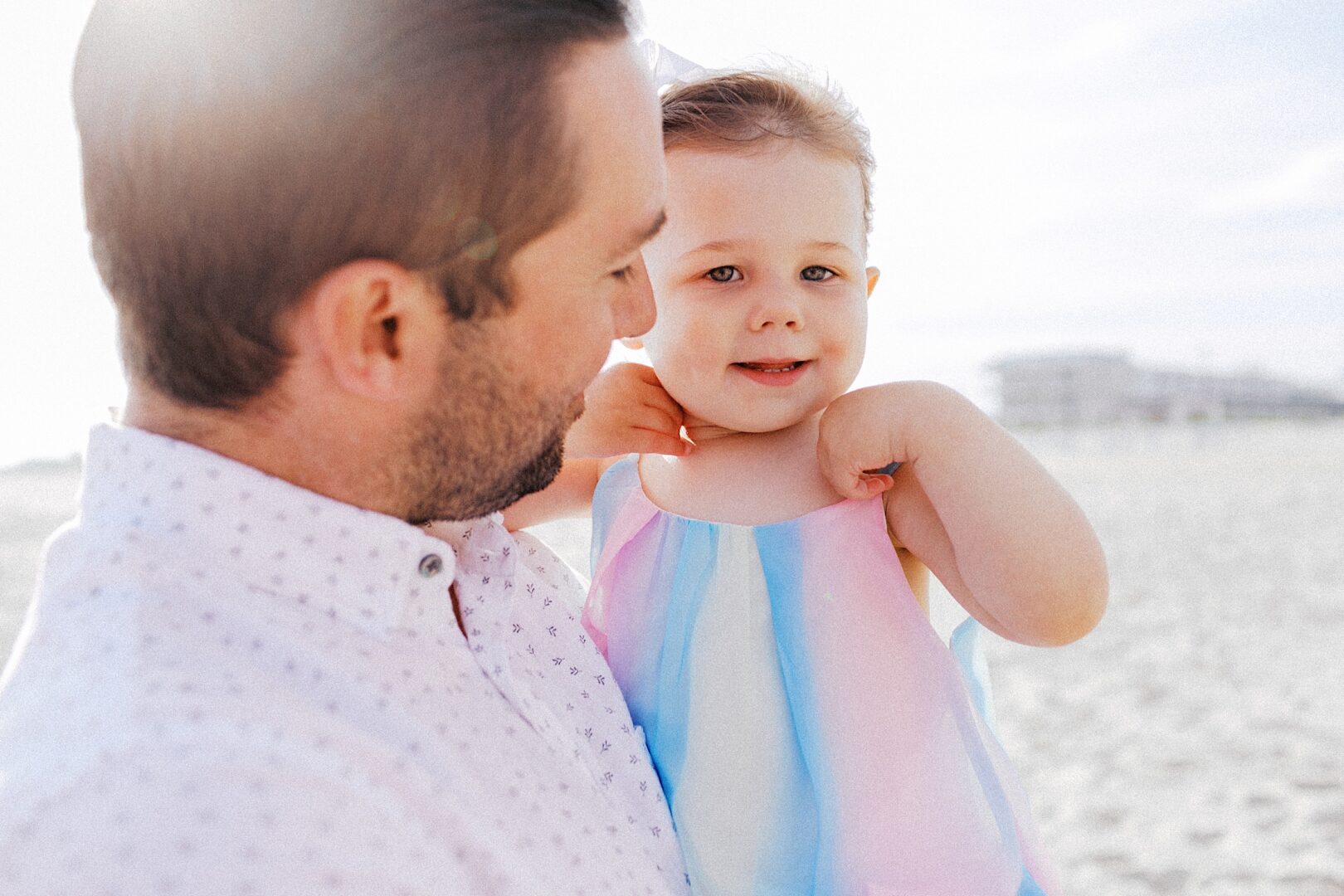 An adult holding a young child at the beach on a sunny day. The child is wearing a pastel-colored dress and looking at the camera.