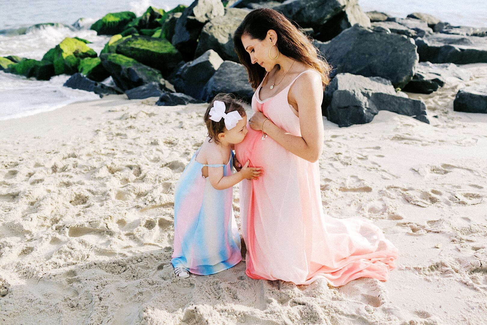 A woman in a pink dress kneels on a sandy beach while a young girl in a blue and pink dress touches her pregnant belly. Large rocks and the ocean are in the background.