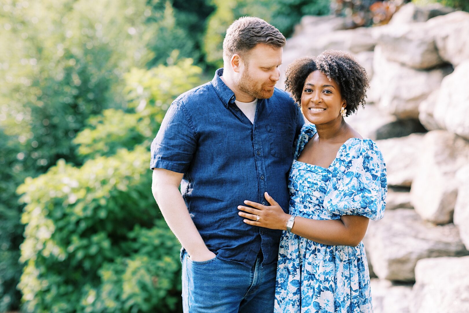 A man and a woman stand close together outdoors by rocks and greenery, both smiling. The man wears a blue shirt and jeans; the woman wears a blue and white floral dress.