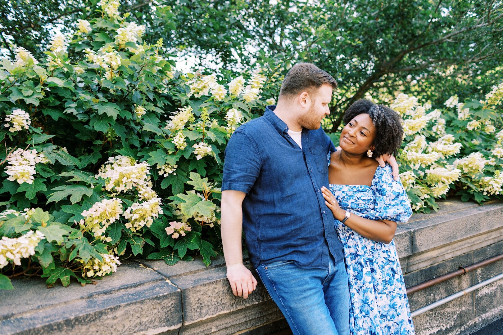 A couple stands together outdoors next to a stone wall and blooming bushes, looking at each other and smiling.