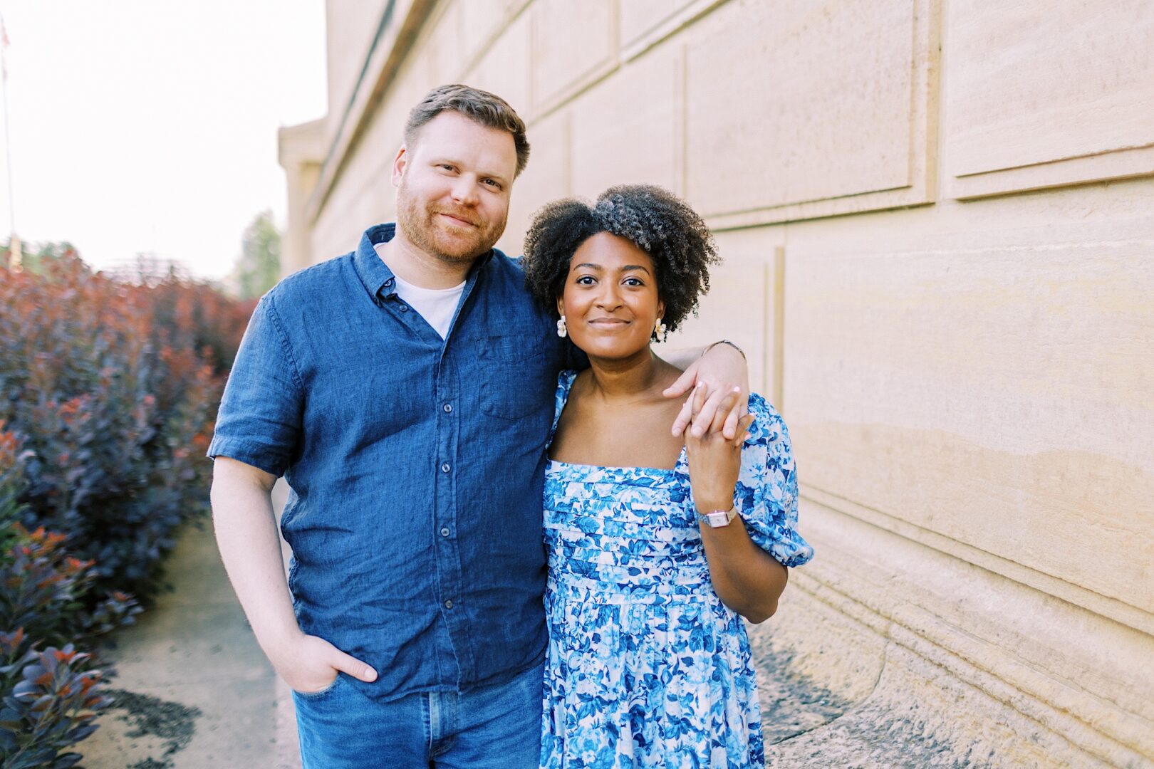 A man and woman stand side by side outdoors, with the man’s arm around the woman’s shoulders. They both smile at the camera, in front of a beige stone wall and shrubbery.