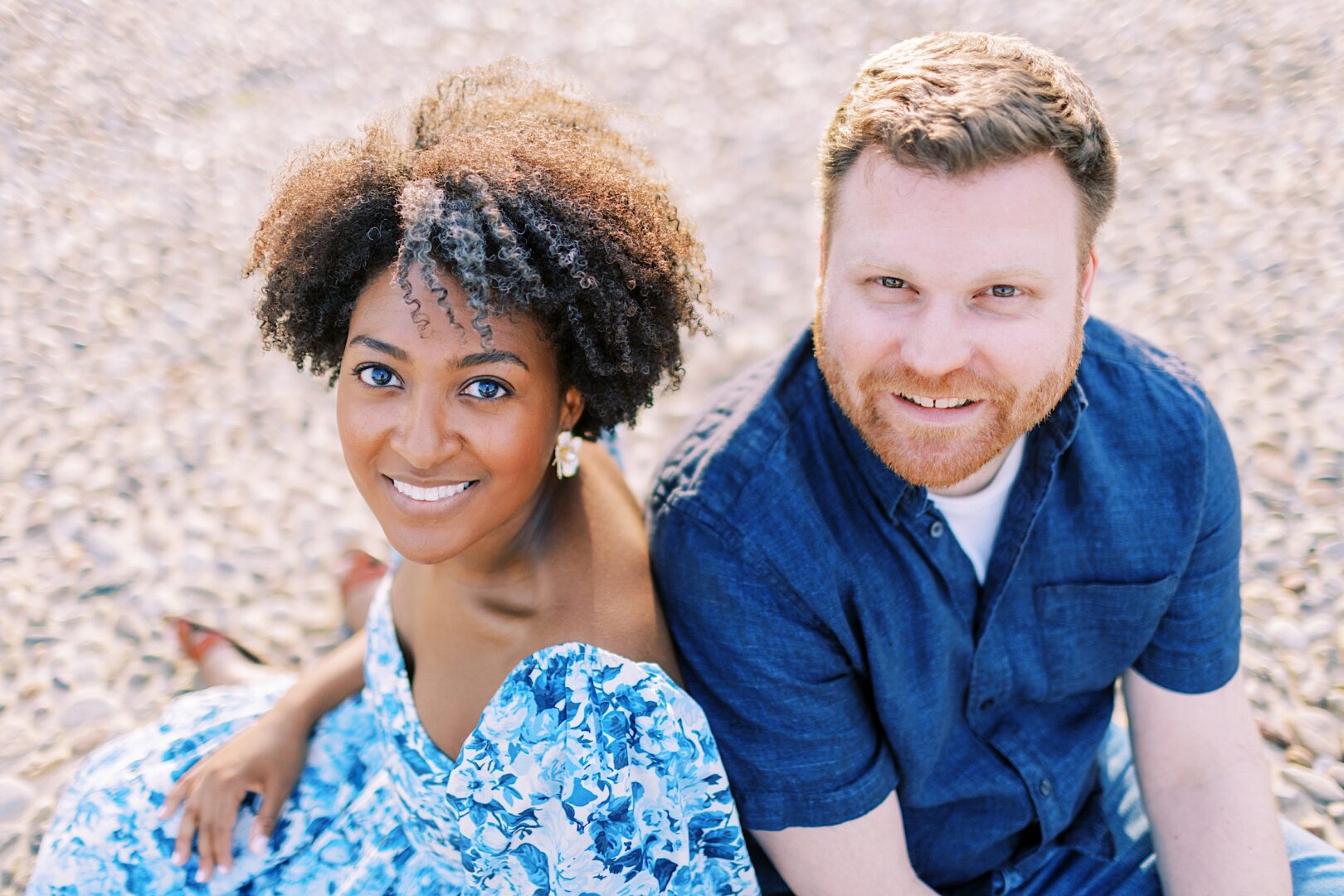 A woman in a blue floral dress and a man in a blue shirt sit next to each other on a sandy surface, both smiling at the camera.