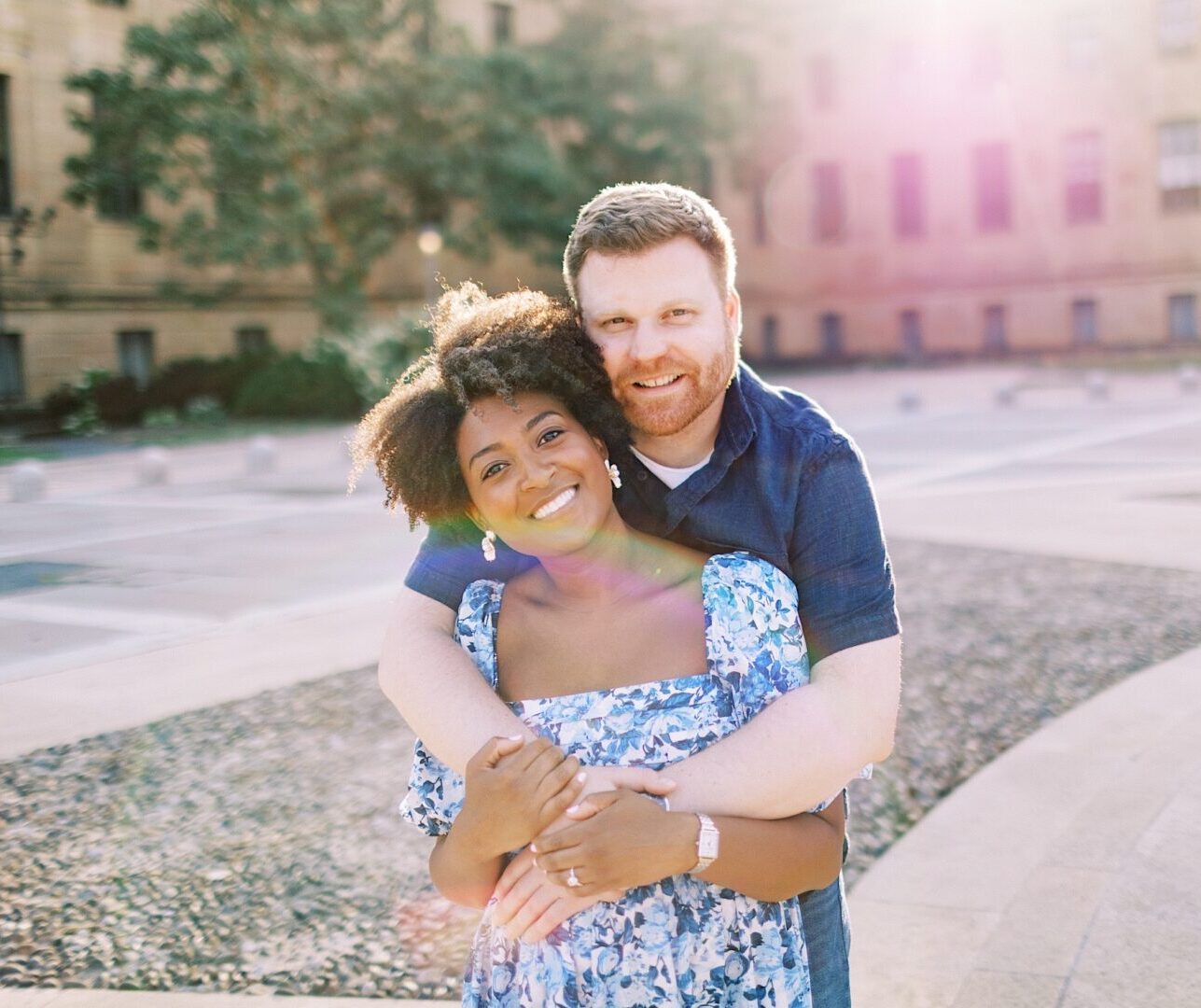 A man stands behind a woman outdoors, hugging her with both arms. They are smiling at the camera in a sunlit, open plaza with a large building in the background.