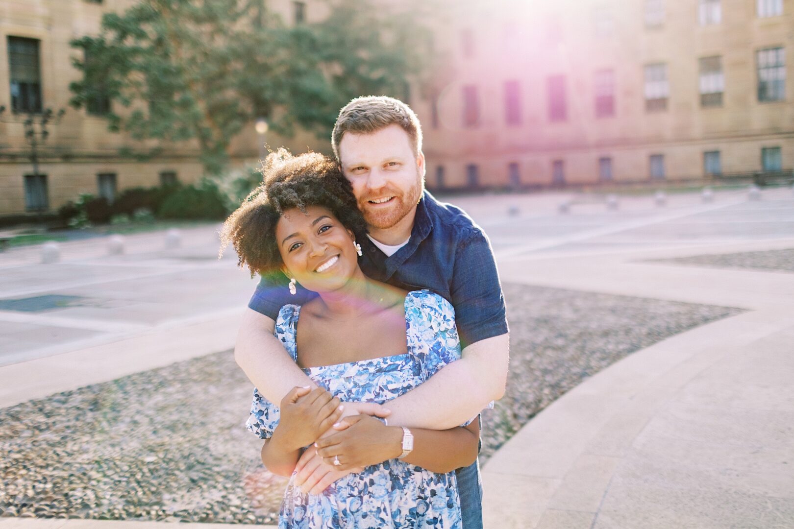 A man stands behind a woman outdoors, hugging her with both arms. They are smiling at the camera in a sunlit, open plaza with a large building in the background.
