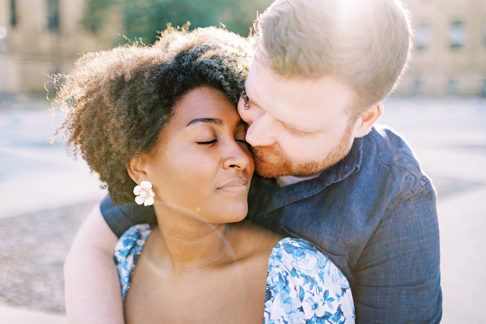 A man embraces a woman from behind outdoors in sunlight. The woman has her eyes closed and wears floral earrings and a blue dress; the man gazes at her.