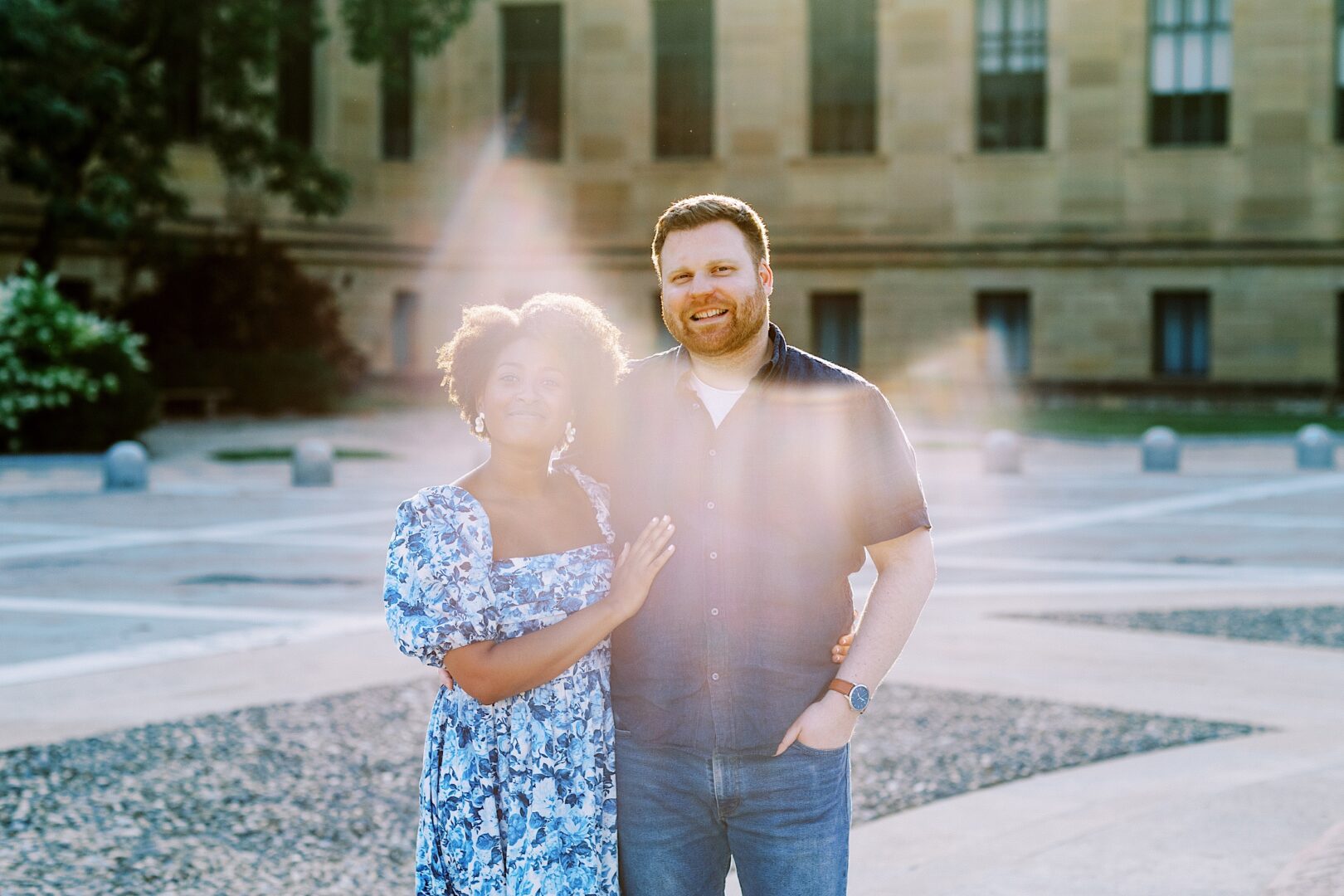 A woman and a man stand together outdoors in sunlight, with the woman’s hand on the man’s chest. A large building is visible in the background.