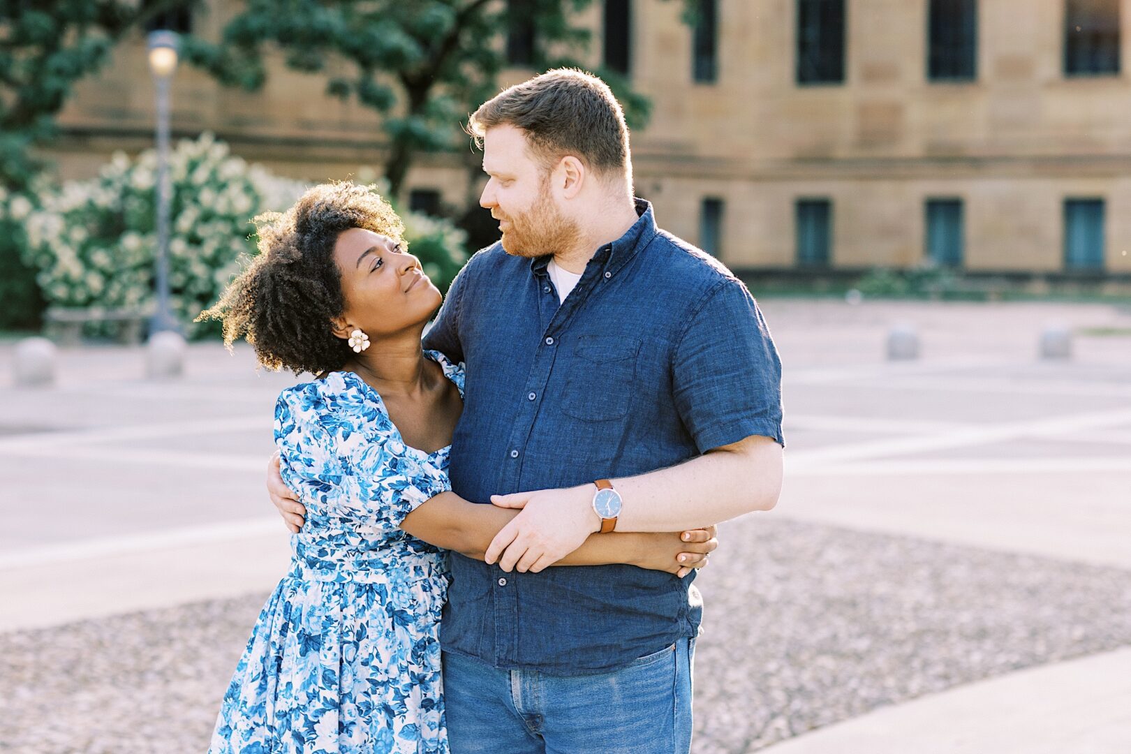 A couple stands outdoors, embracing and looking at each other, with trees and a large building in the background.