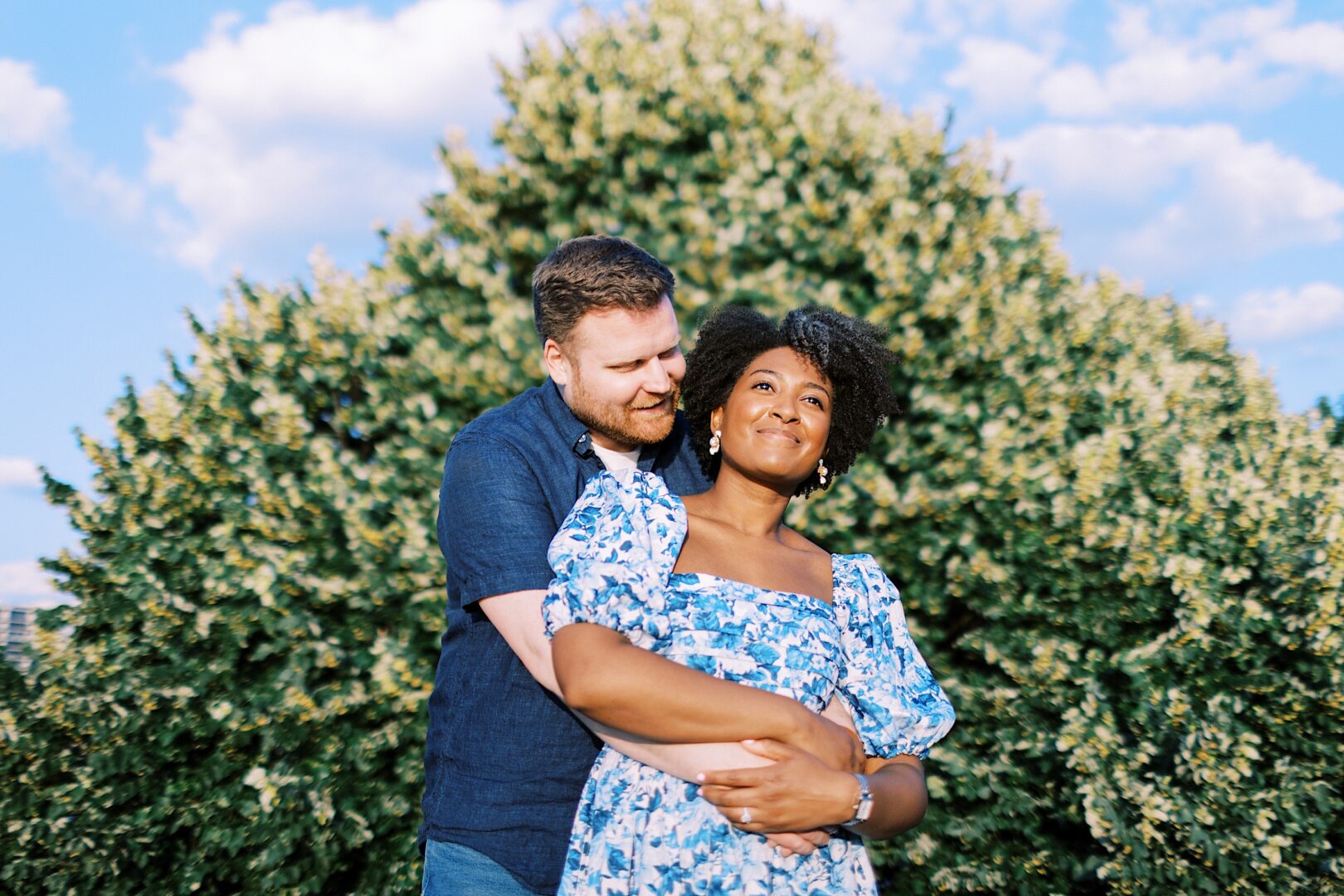 A man stands behind a woman, embracing her as they both smile. They are outdoors in front of a large leafy tree under a blue sky with scattered clouds.