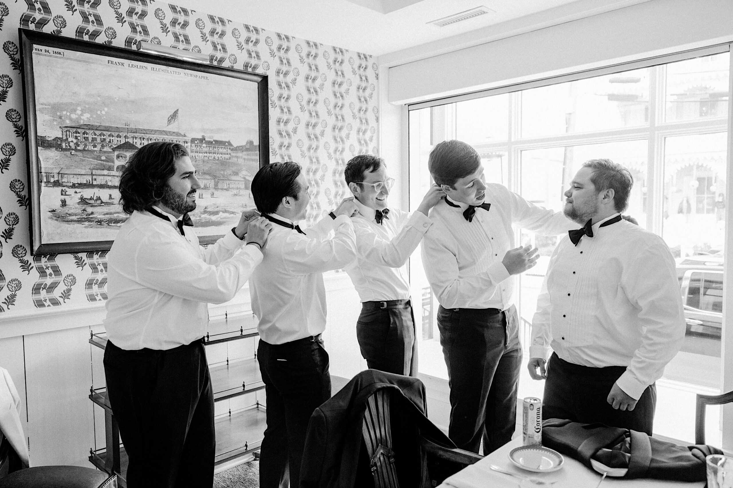 Five men in white dress shirts and bow ties stand together in a bright room at a New Jersey Beach Wedding Venue, helping each other adjust their collars and ties.