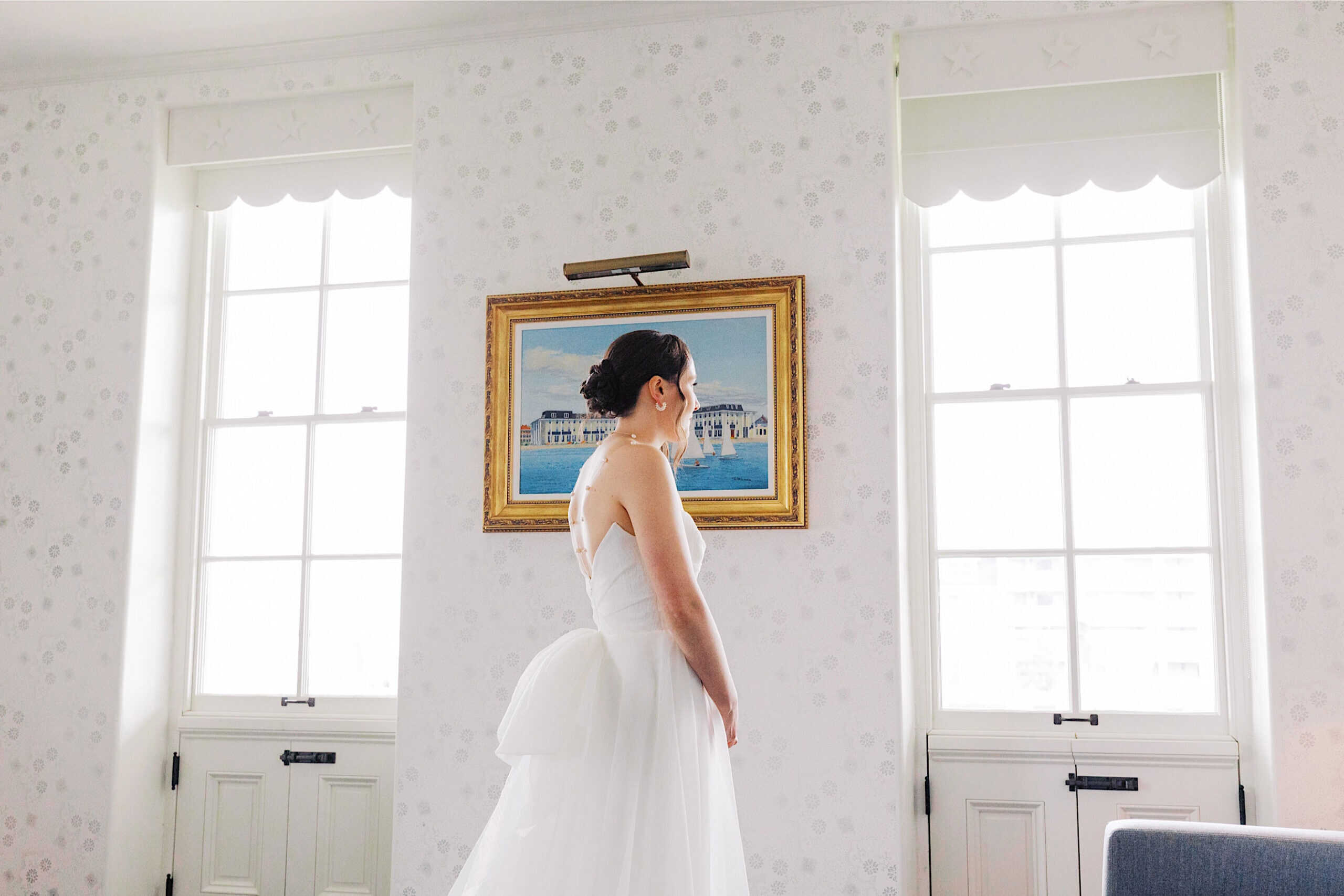 A woman in a white strapless wedding dress stands indoors between two tall windows, facing sideways, with a framed painting on the wall behind her—capturing the elegance of a New Jersey Beach Wedding Venue.