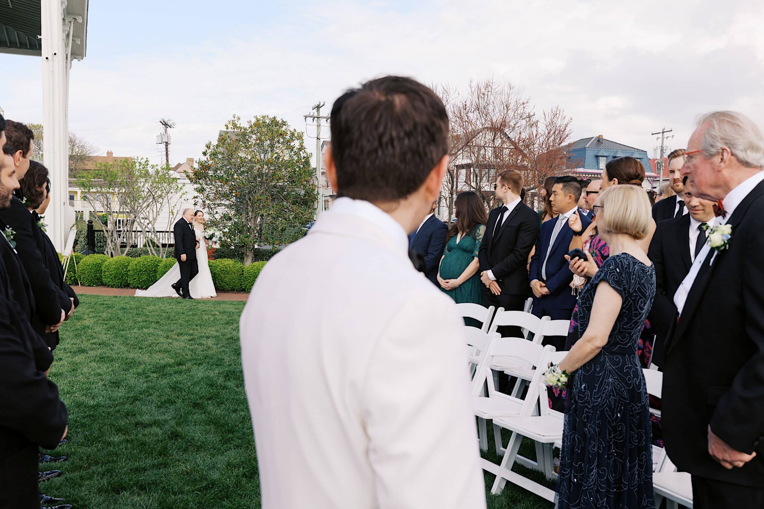 A bride walks down the aisle outdoors at a New Jersey Beach Wedding Venue as guests and a person in a white suit, seen from behind, watch during the ceremony.