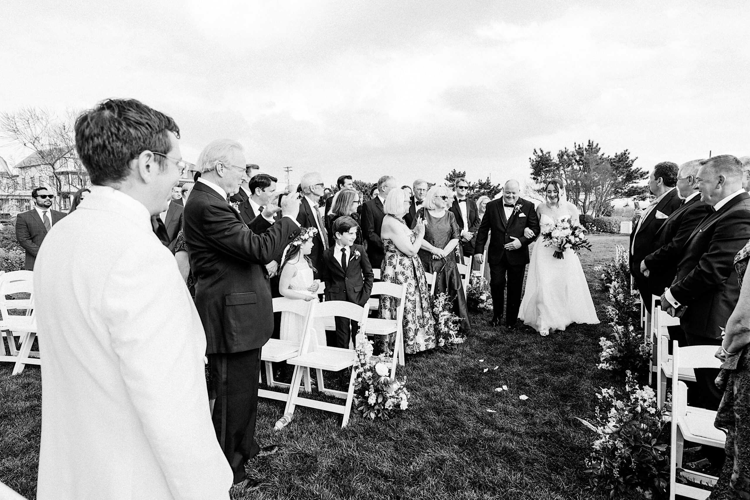 A bride walks down an outdoor aisle at a New Jersey Beach Wedding Venue with an older man, while guests on both sides watch and take photos; the groom stands at the front in a white jacket.