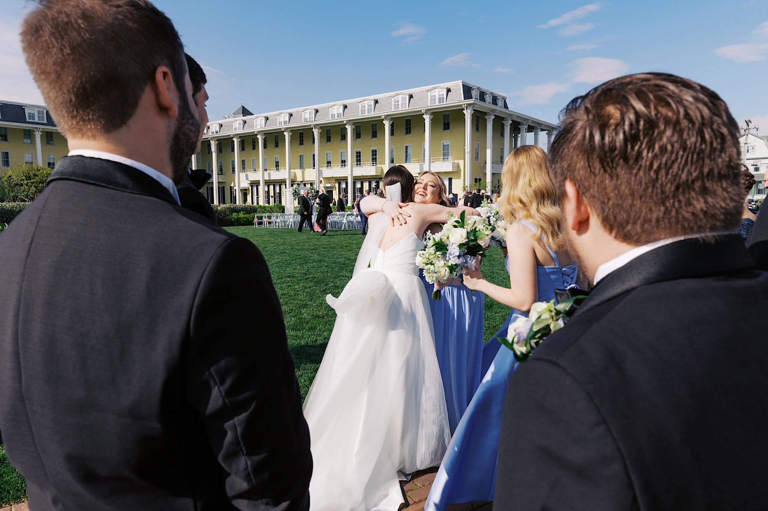 Two women in formal dresses hug outdoors at a New Jersey Beach Wedding Venue, surrounded by guests in suits and gowns, with a large yellow building in the background.