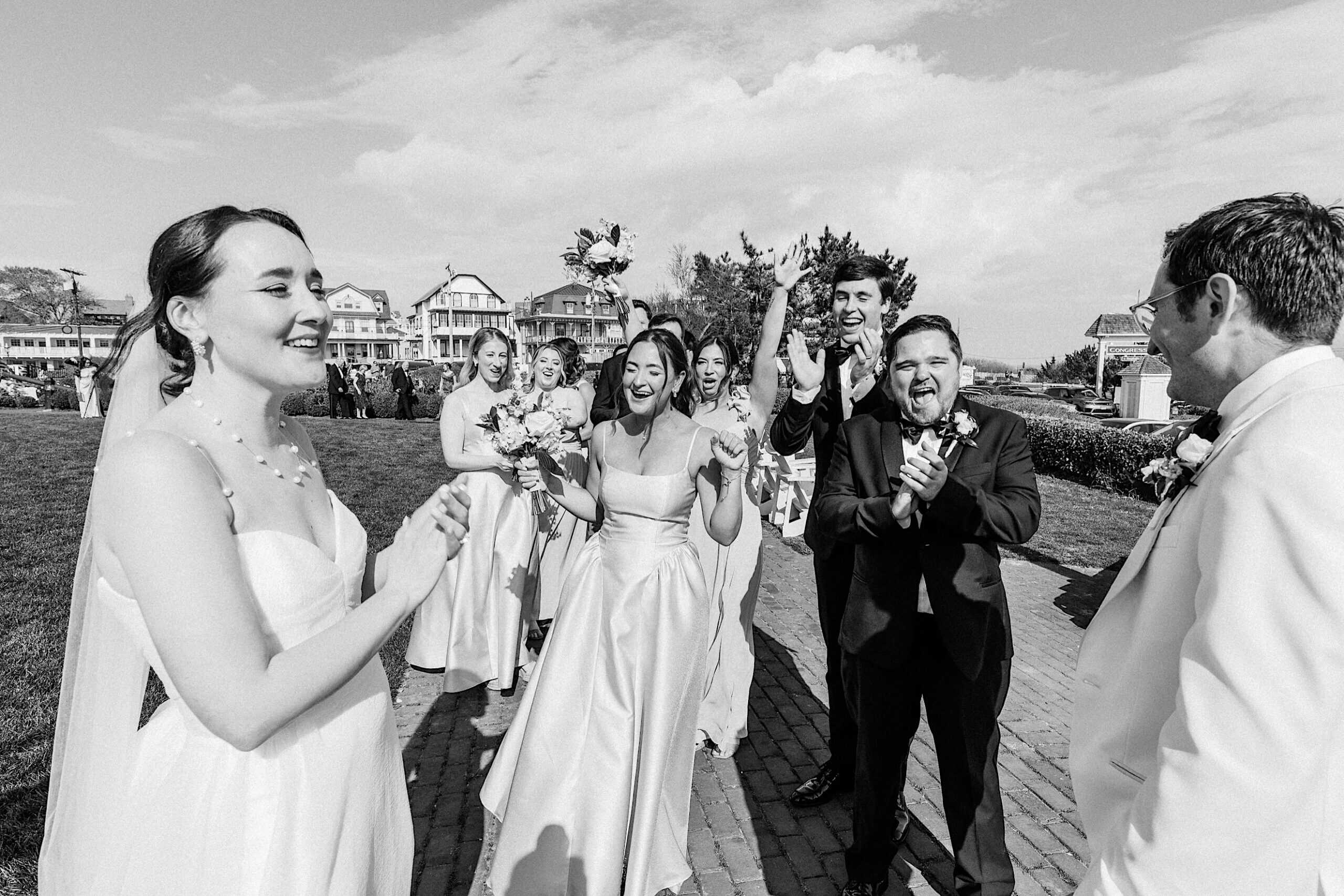 A bride and groom celebrate outdoors with their wedding party, who clap and cheer. The group is dressed formally and appears joyful on a sunny day at a stunning New Jersey Beach Wedding Venue.