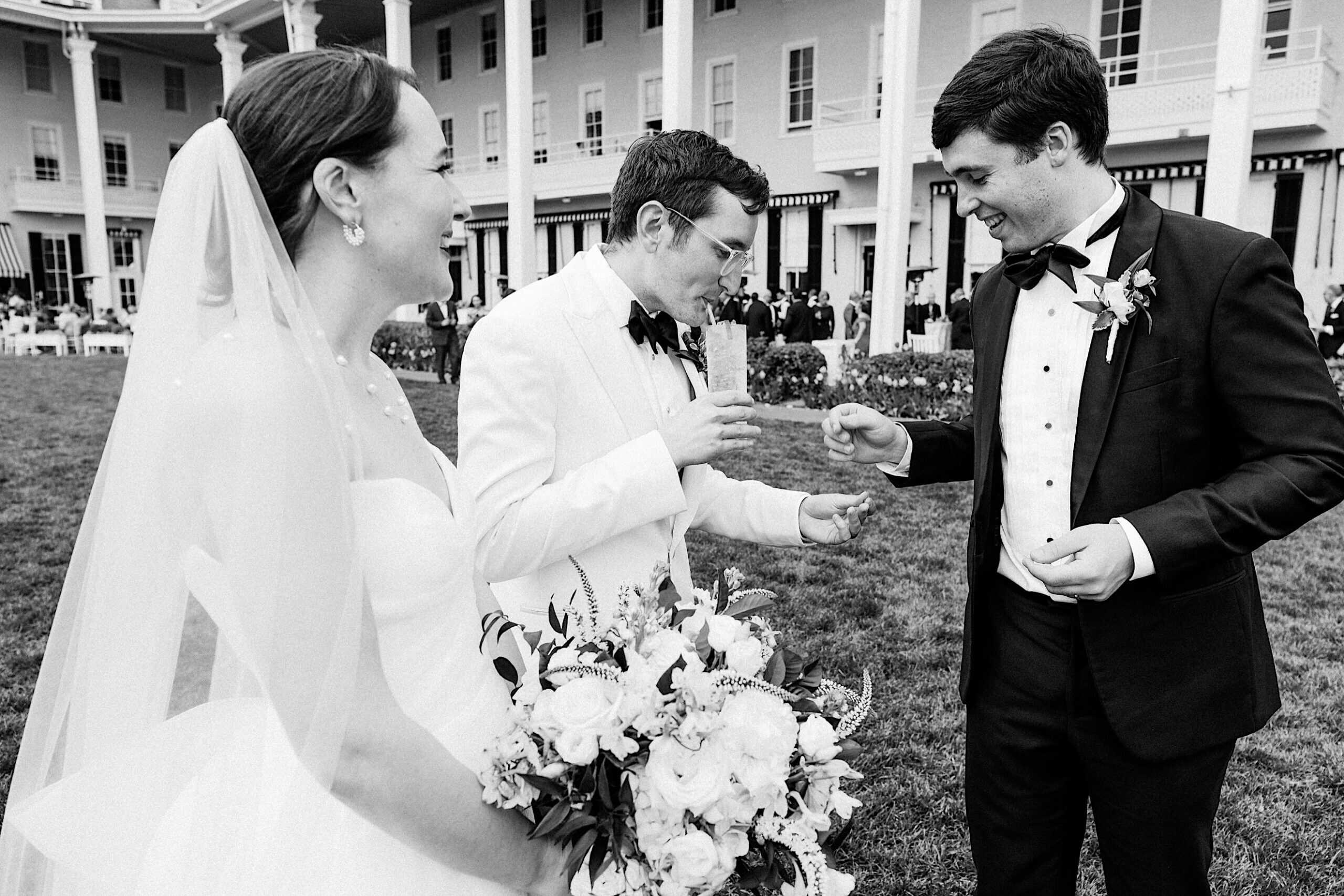 A bride holding a bouquet stands beside two men in tuxedos, one sipping from a glass, outdoors near a large building at a New Jersey Beach Wedding Venue during a formal event.