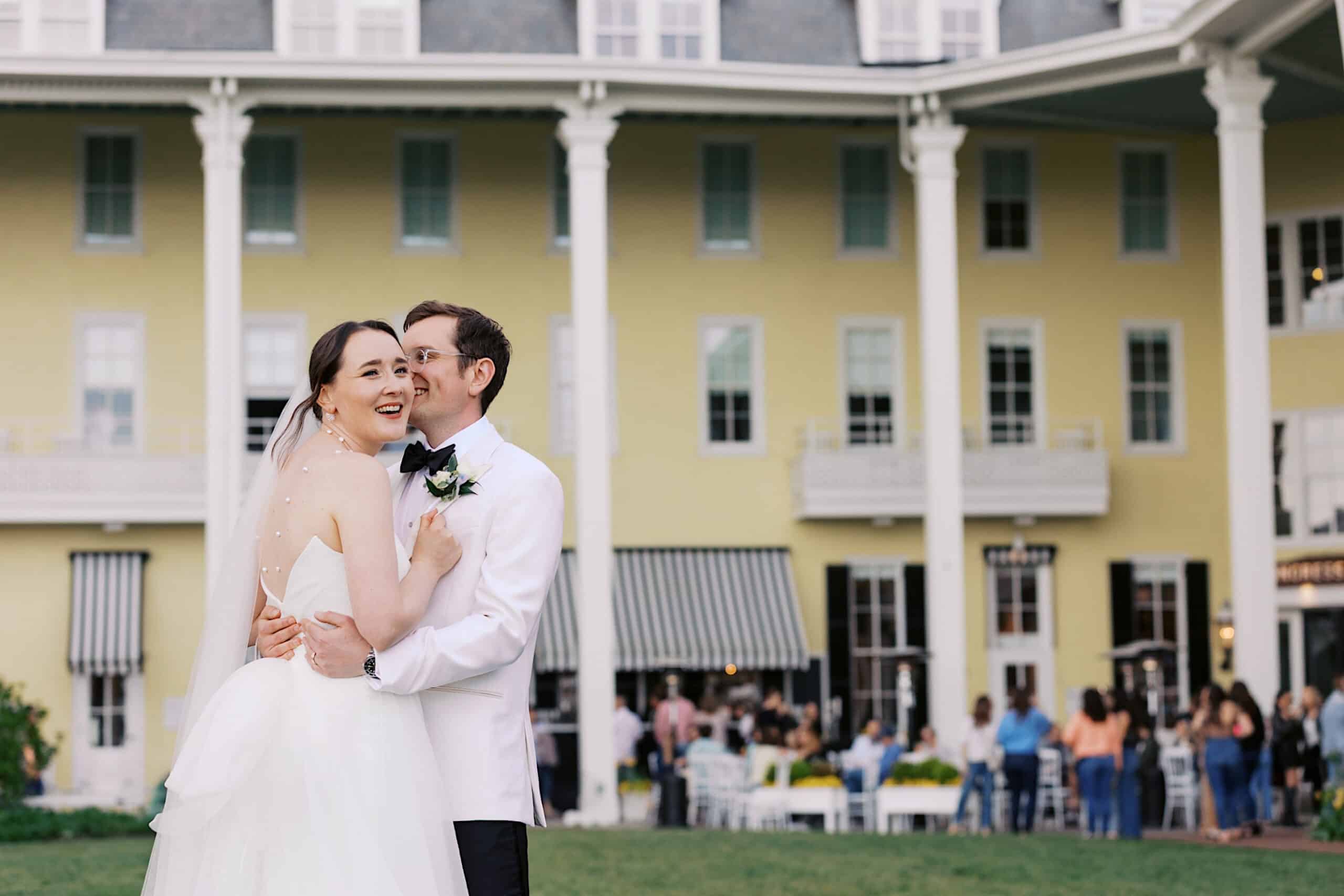 A bride and groom embrace and smile outside a large yellow building with columns, as guests gather in the background at this charming New Jersey Beach Wedding Venue.
