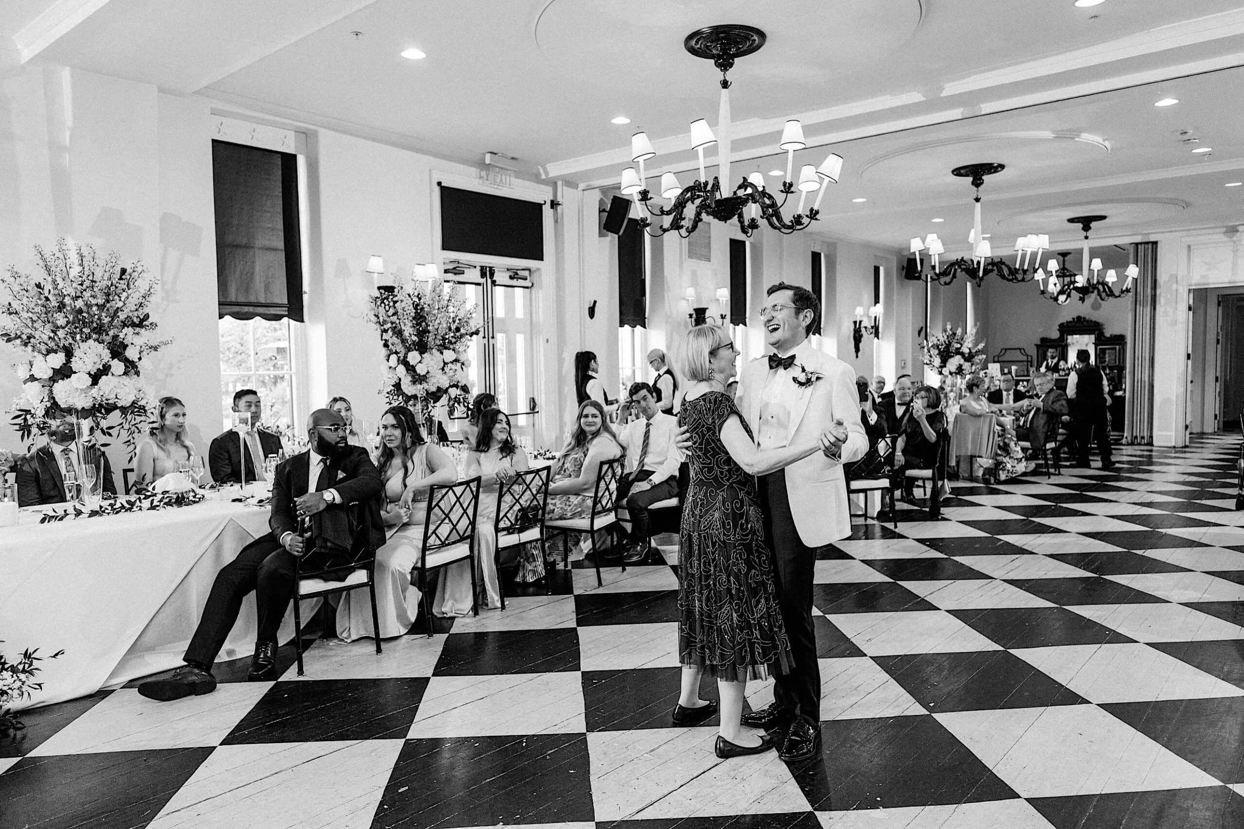 A groom in a light suit dances with an older woman on a checkered floor, while seated guests watch in a decorated reception hall at a charming New Jersey Beach Wedding Venue.
