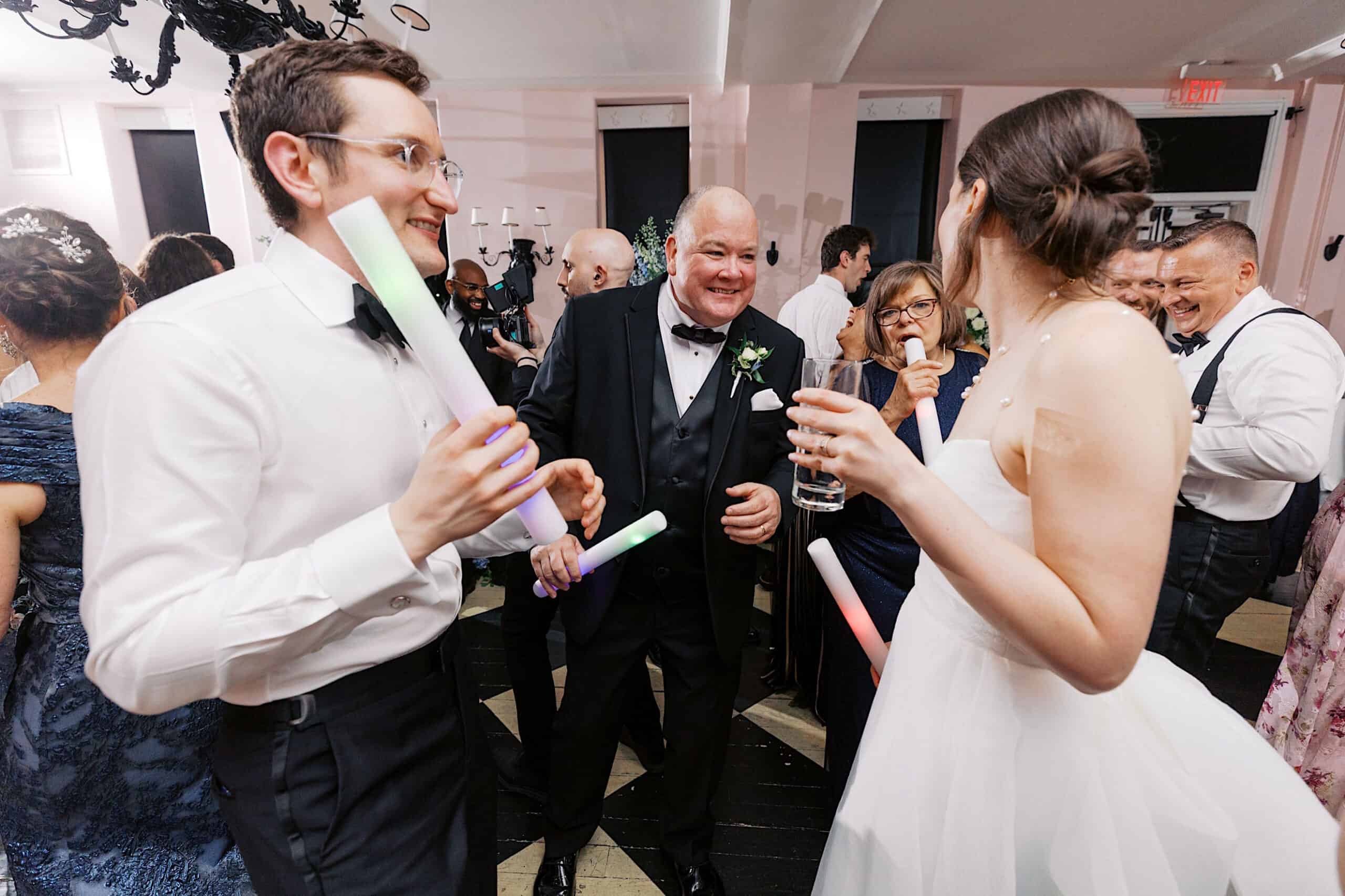 A group of people in formal attire, including a bride and groom, are smiling and holding glow sticks at a lively indoor party or wedding reception at a New Jersey Beach Wedding Venue.