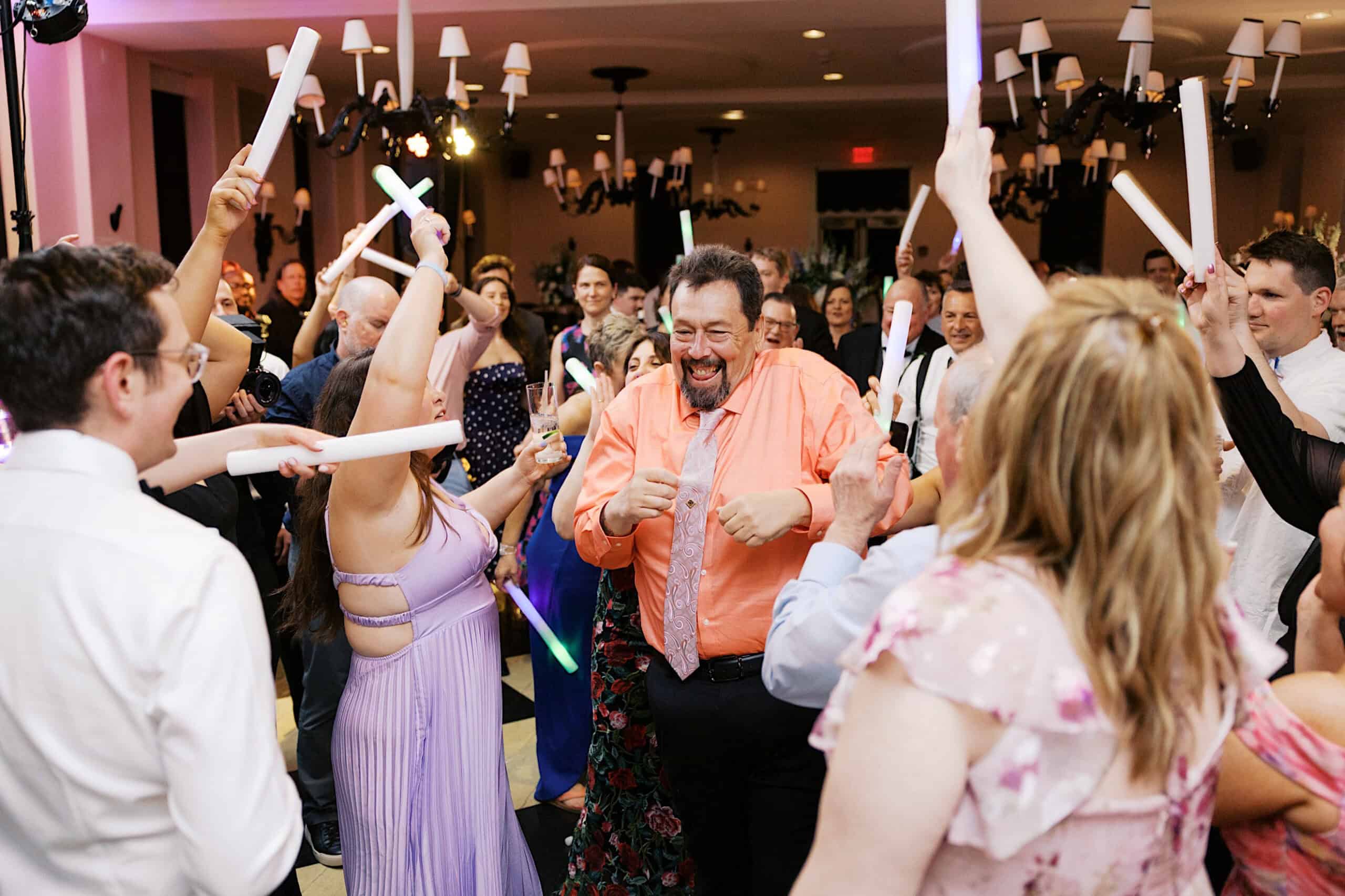 A group of people dance and celebrate indoors, holding glowing light sticks, with a man in an orange shirt smiling at the center—capturing the vibrant joy of a New Jersey Beach Wedding Venue celebration.