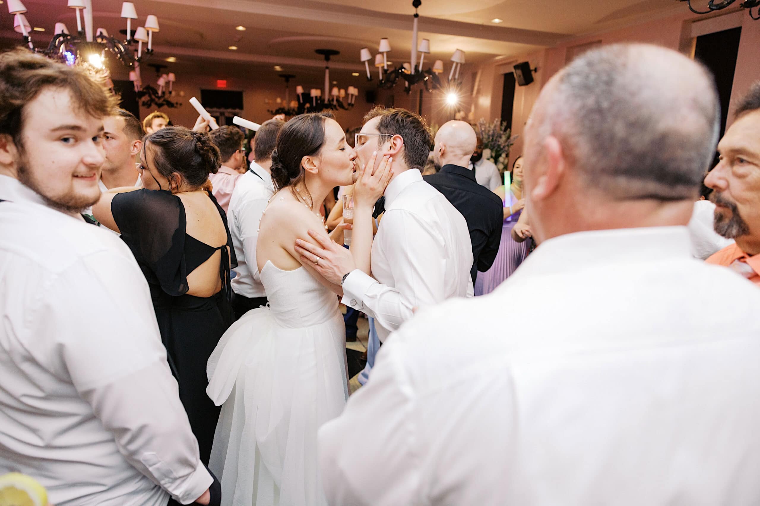 A bride and groom kiss in the middle of a crowded wedding reception at a stunning New Jersey Beach Wedding Venue, surrounded by guests dressed in formal attire.