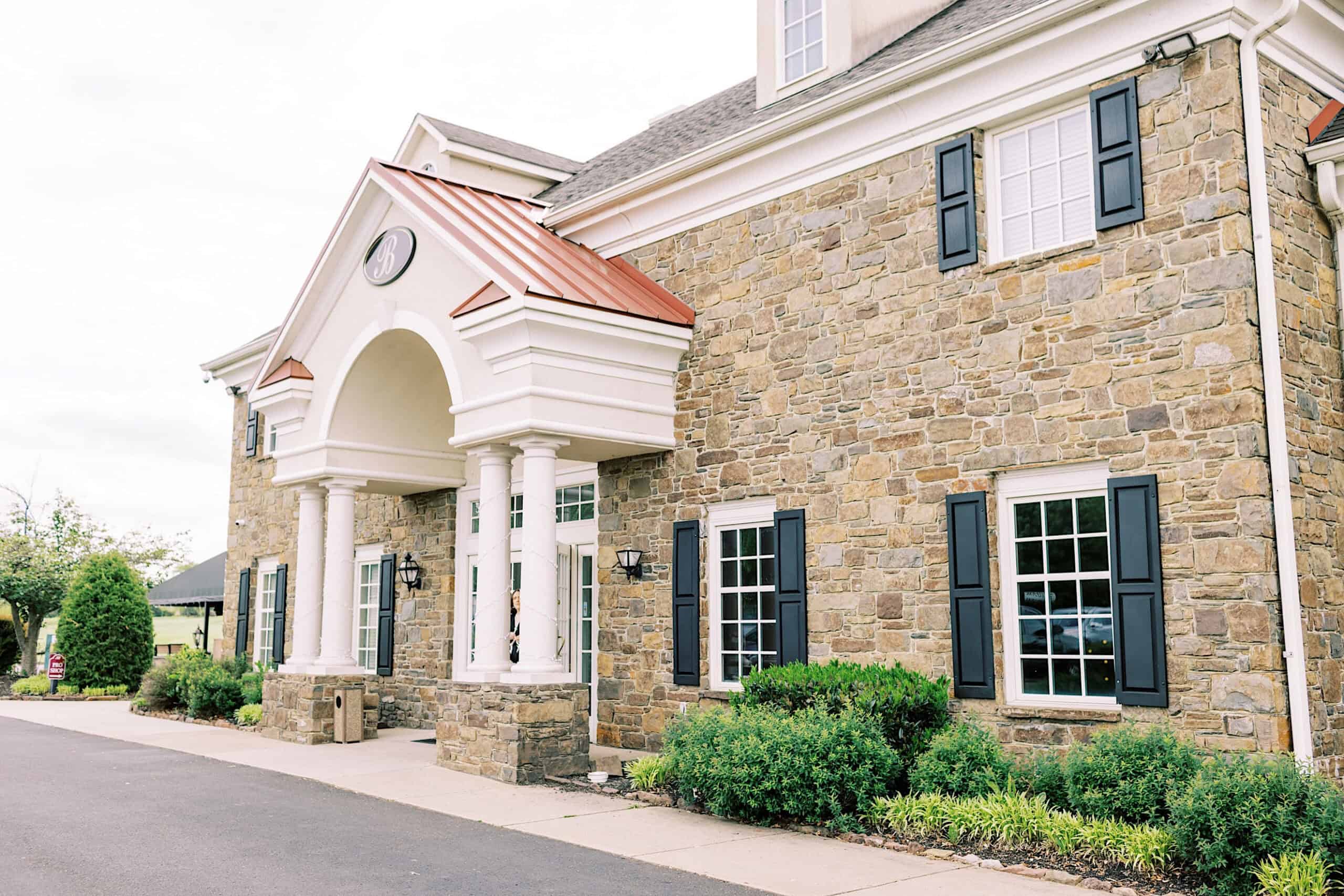 A two-story stone building with white columns, black shutters, landscaped bushes, and a circular driveway under a partly cloudy sky—perfect for a Great Gatsby Sweet 16 celebration.