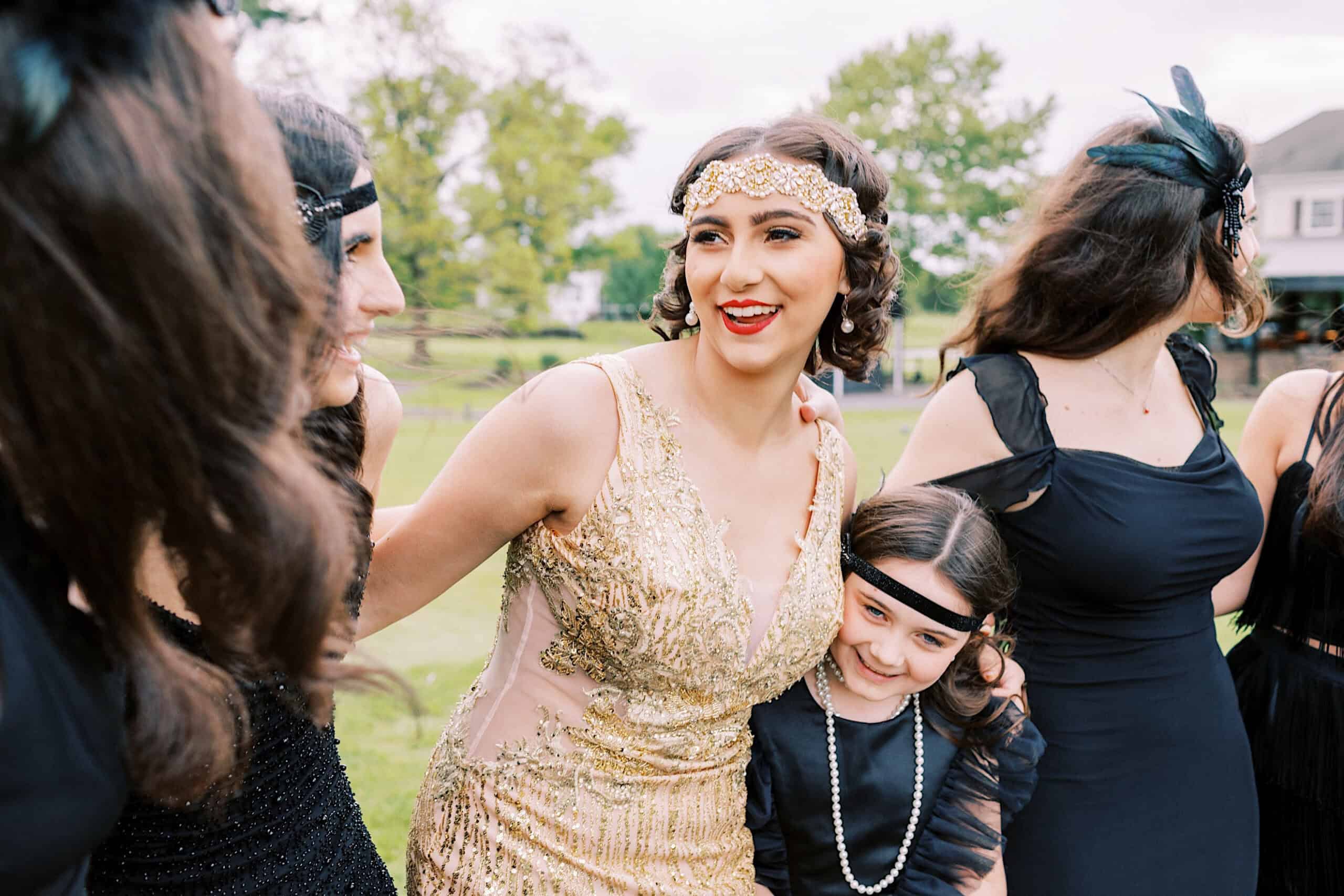 A group of women and a young girl in 1920s-style outfits celebrate a Great Gatsby Sweet 16, with one woman in a gold dress, all standing outdoors and smiling.