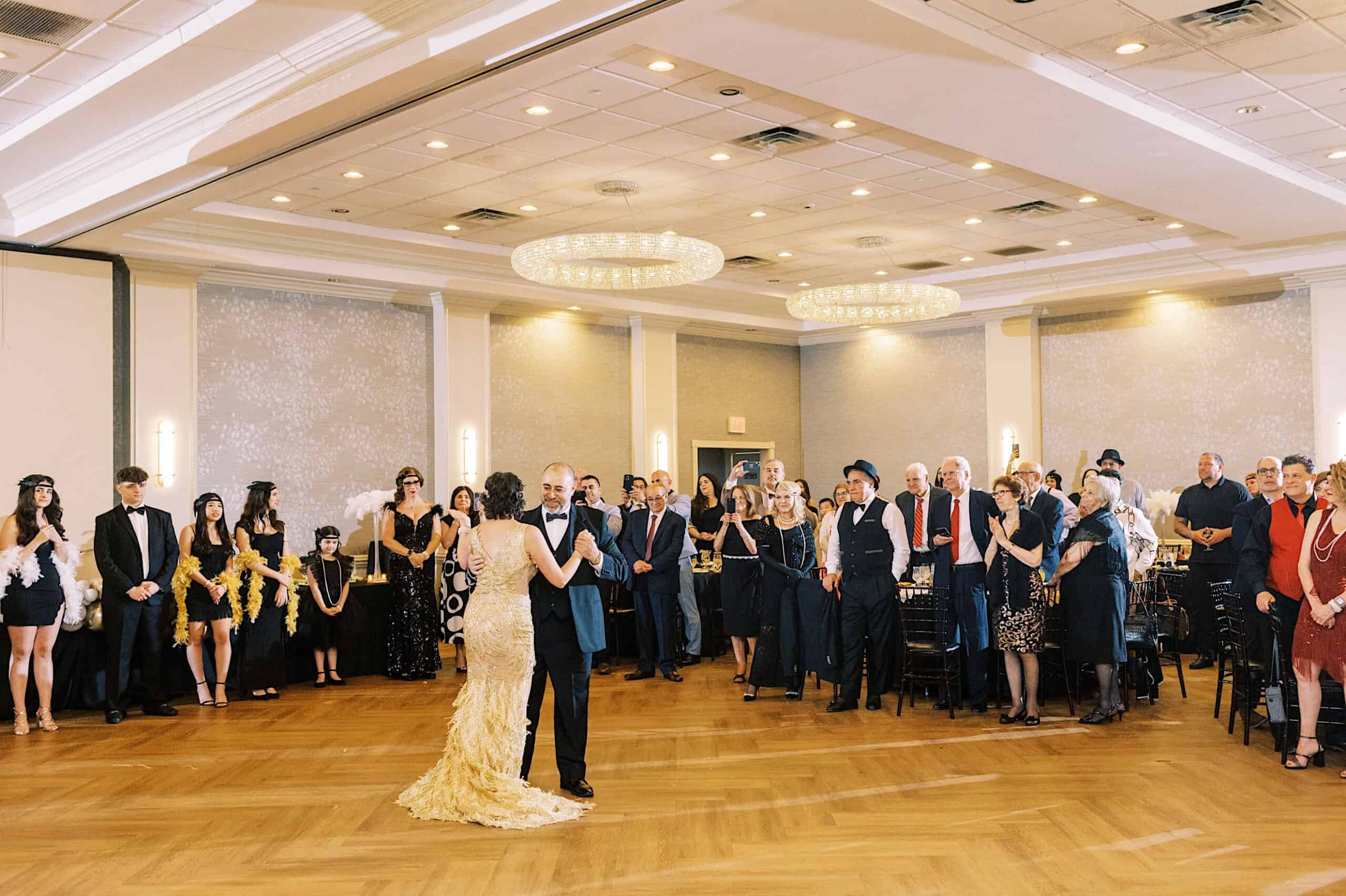 A couple dances in the center of a ballroom at a glamorous Great Gatsby Sweet 16, while a large group of guests in formal attire watches and stands around the room.