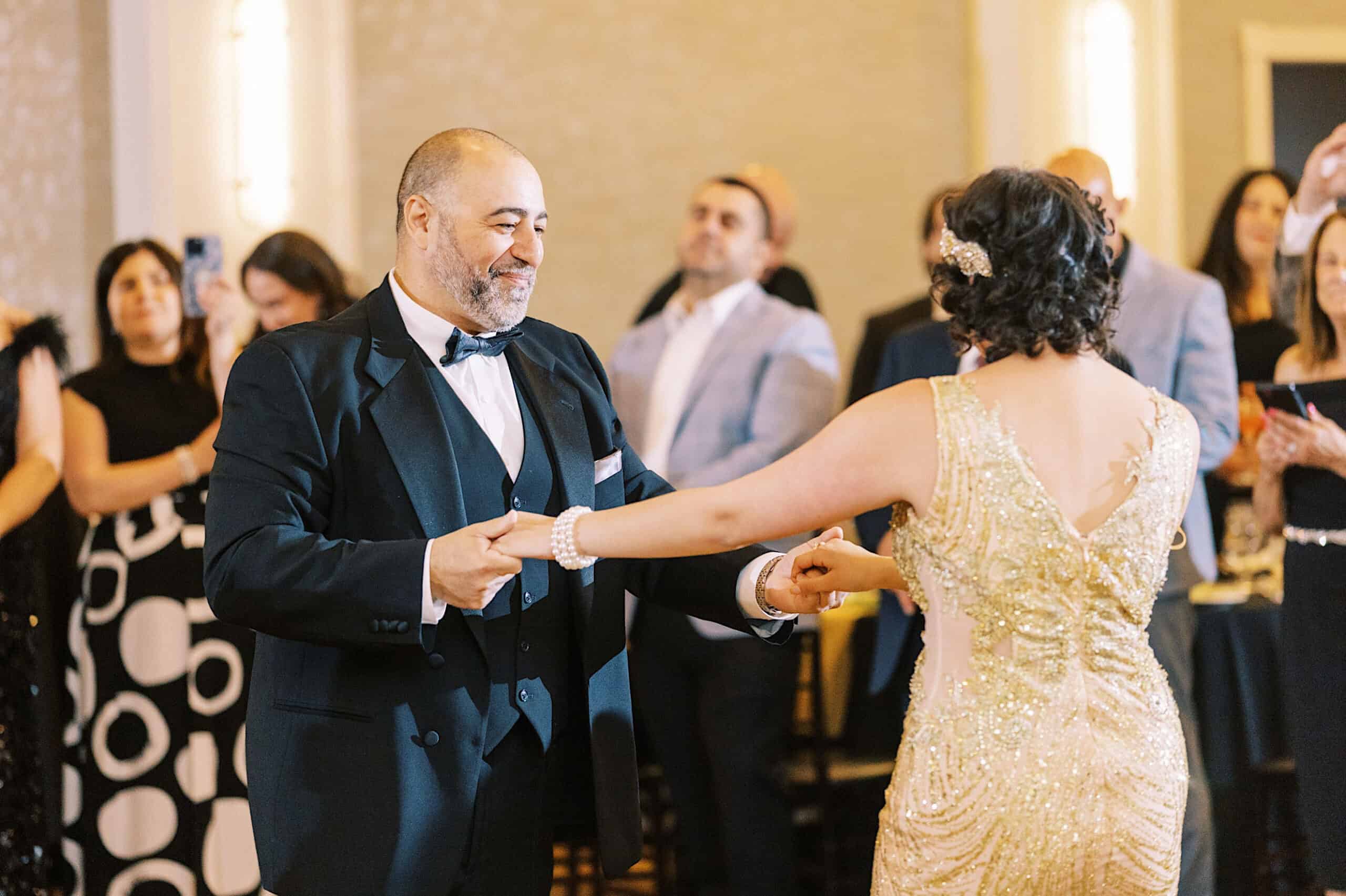 A man in a tuxedo dances with a woman in a gold dress at a Great Gatsby Sweet 16, while guests watch in the background.