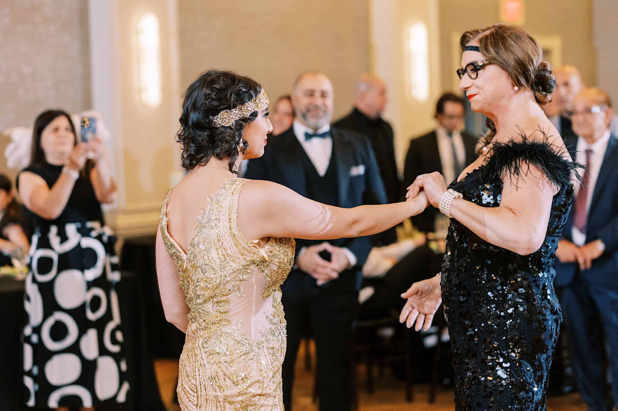 Two women in formal dresses dance together at a Great Gatsby Sweet 16, while people in the background watch and take photos.