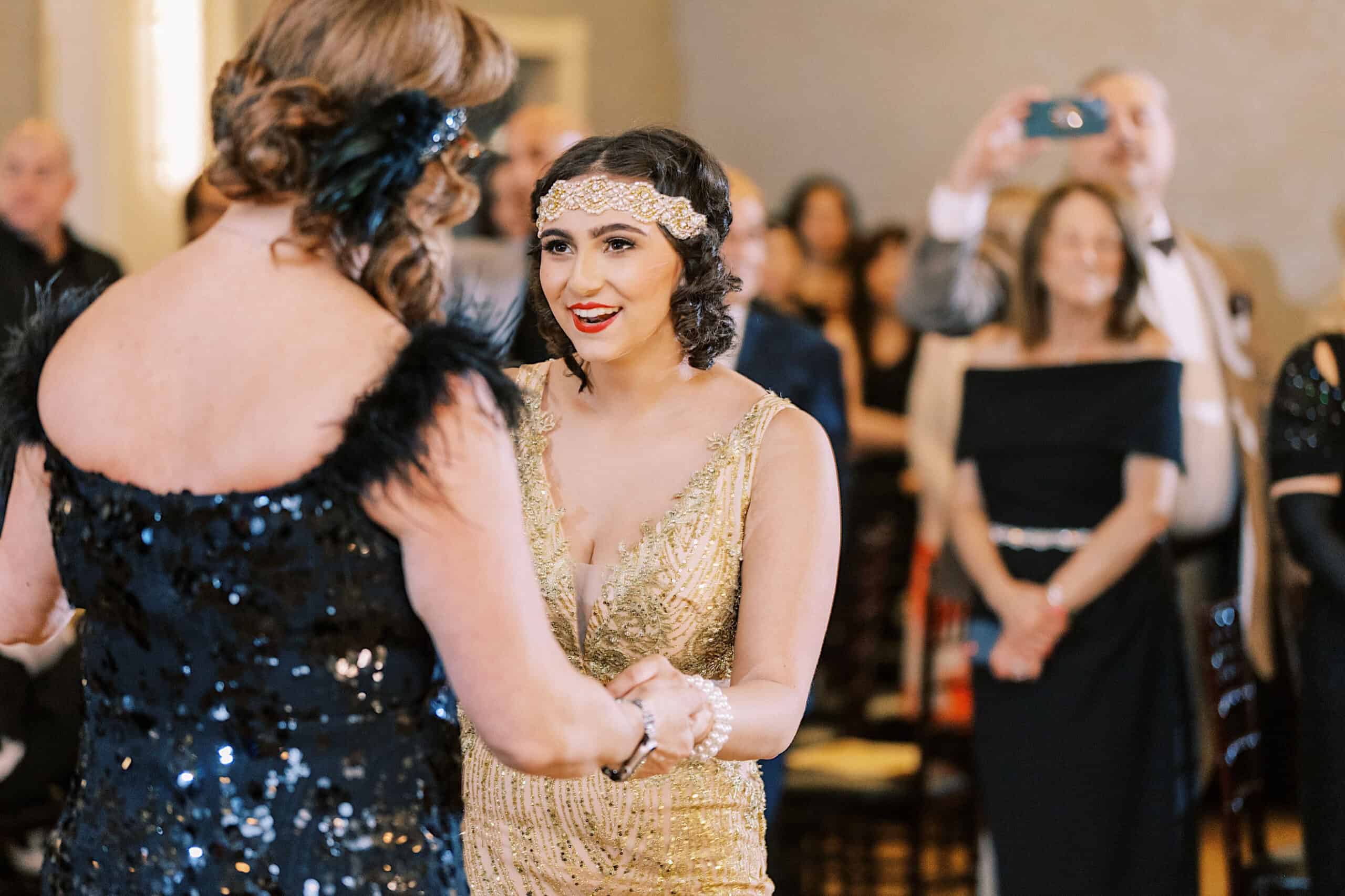 Two women in formal attire, one in gold and one in black sequins, dance together at a Great Gatsby Sweet 16 as people watch and take photos in the background.