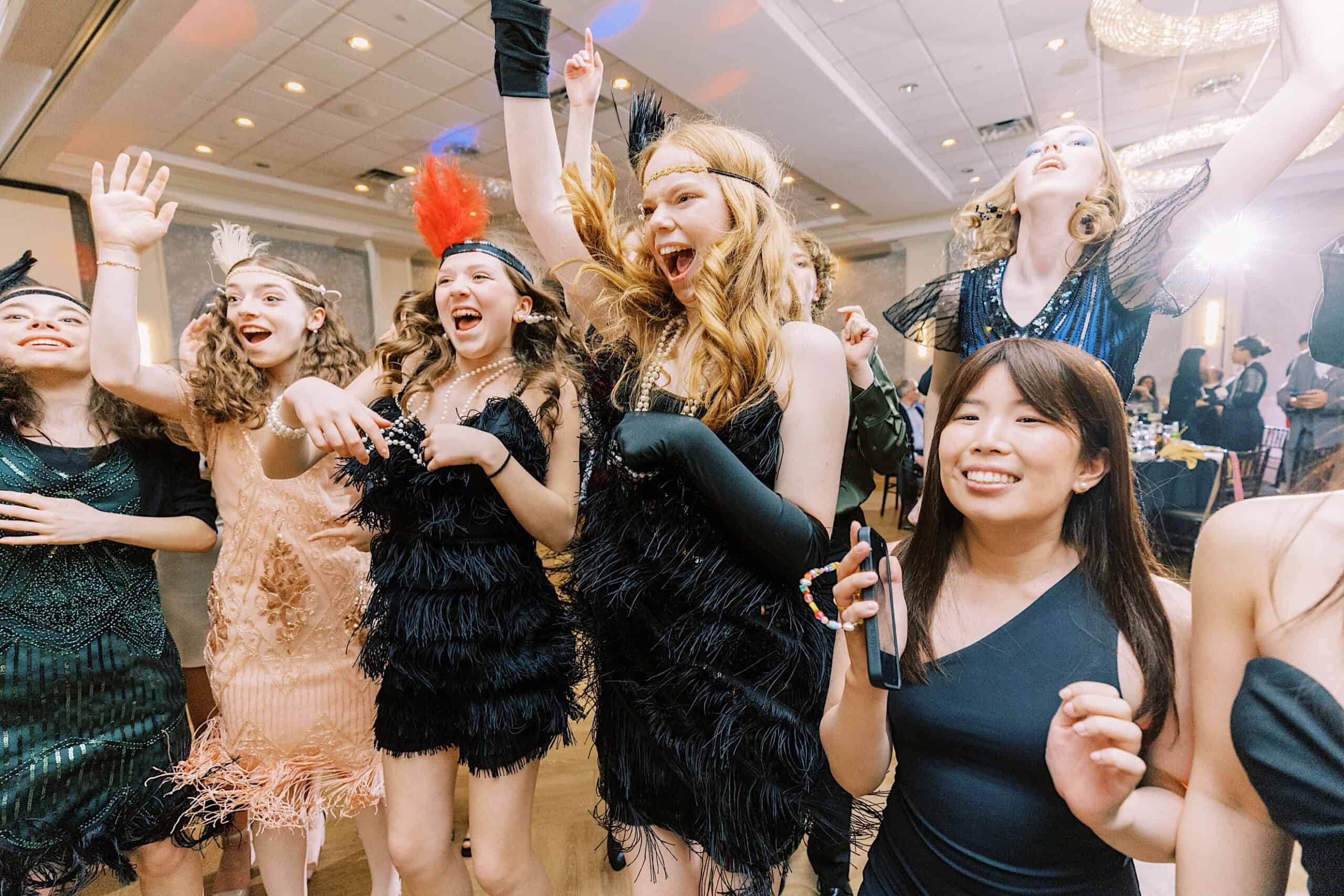 A group of young women in flapper-style dresses and headbands enthusiastically celebrate and dance at a glamorous Great Gatsby Sweet 16 indoor event.