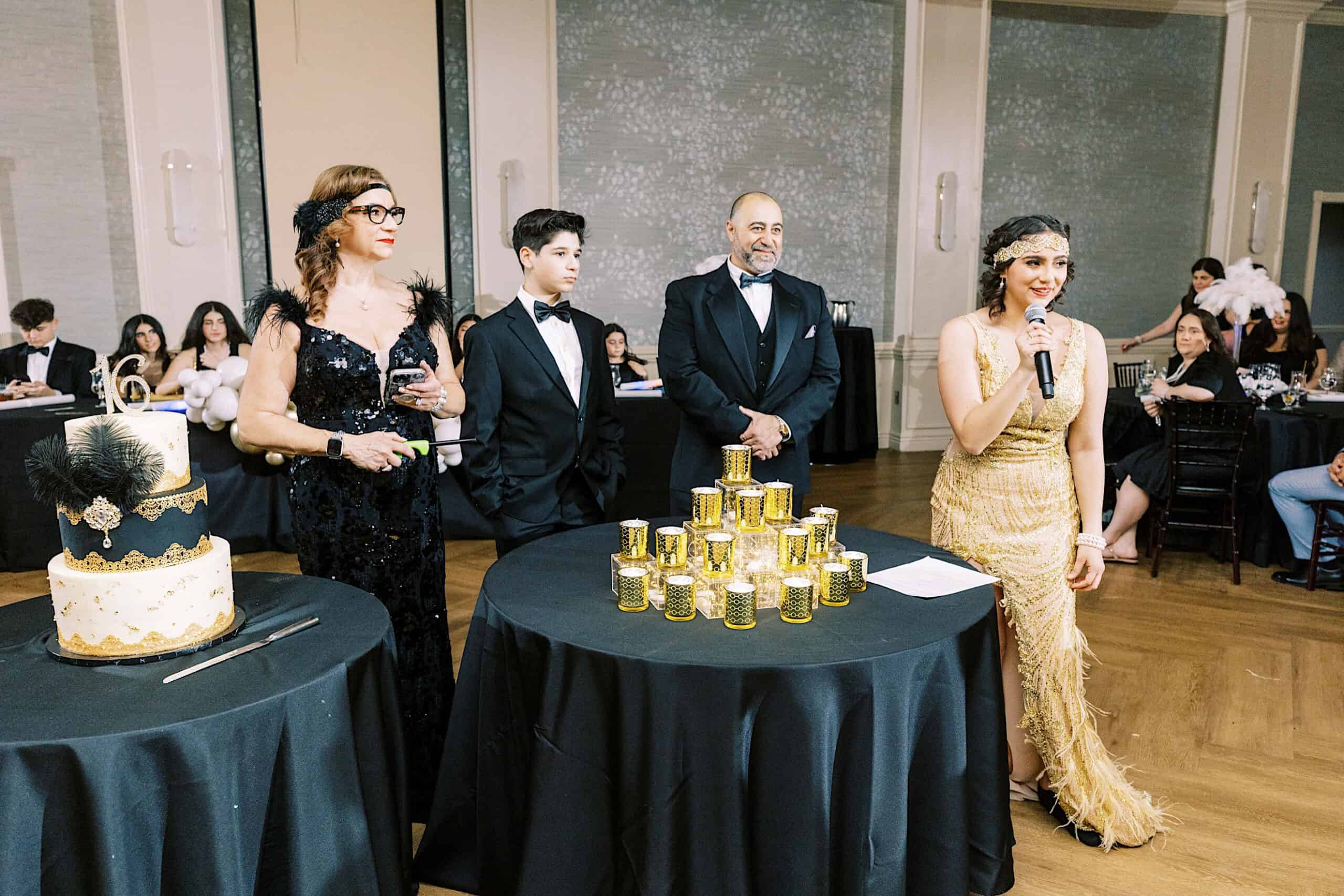 A woman in a gold dress speaks into a microphone beside a table with stacked gold candles; three people in formal attire stand nearby at an elegant Great Gatsby Sweet 16 event.