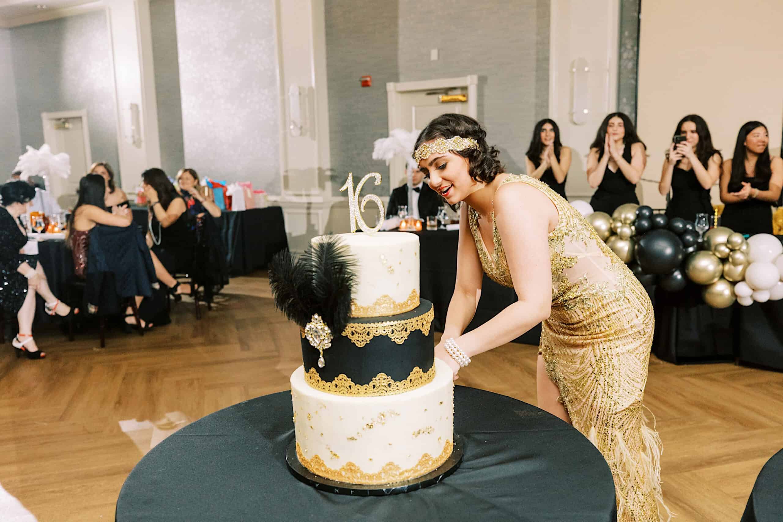 A young woman in a gold dress cuts a two-tiered cake at her Great Gatsby Sweet 16, surrounded by black and gold decorations and guests seated and standing in the background.