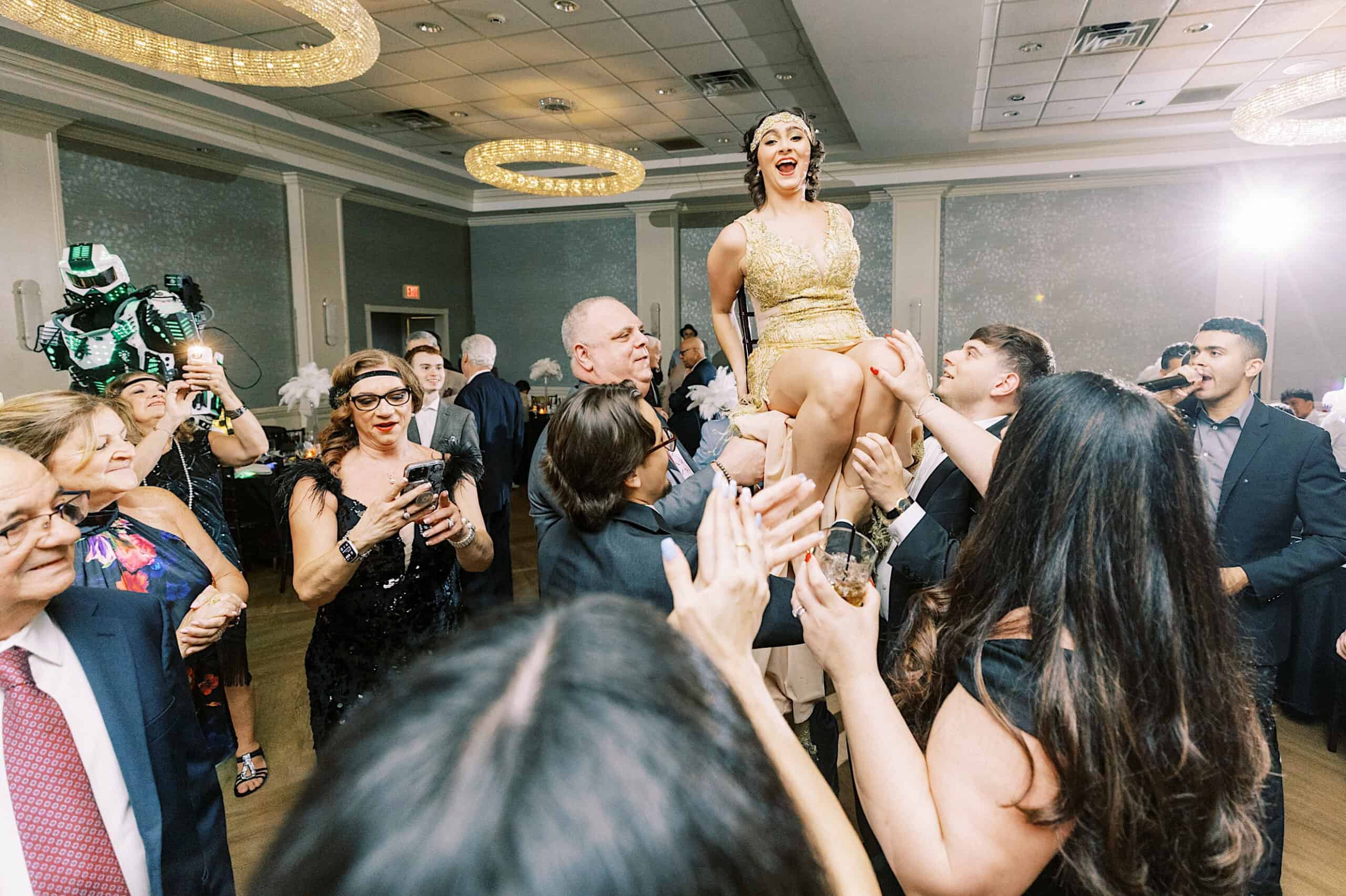 At a Great Gatsby Sweet 16, a woman in a gold dress is lifted on a chair in a crowded, festive banquet hall while guests cheer and take photos.