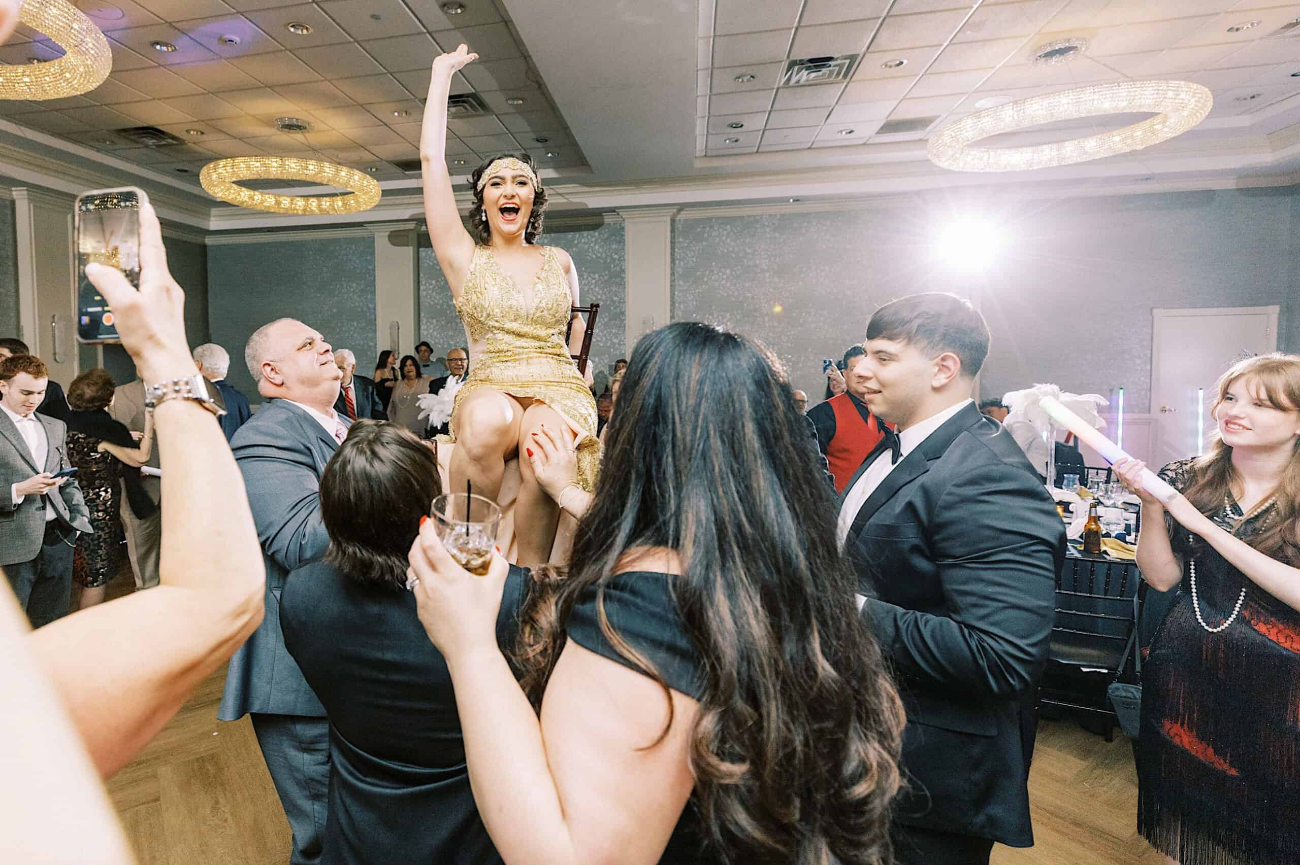 A woman in a gold dress is lifted on a chair by guests at a glamorous Great Gatsby Sweet 16, with people around her smiling and raising glasses in celebration.