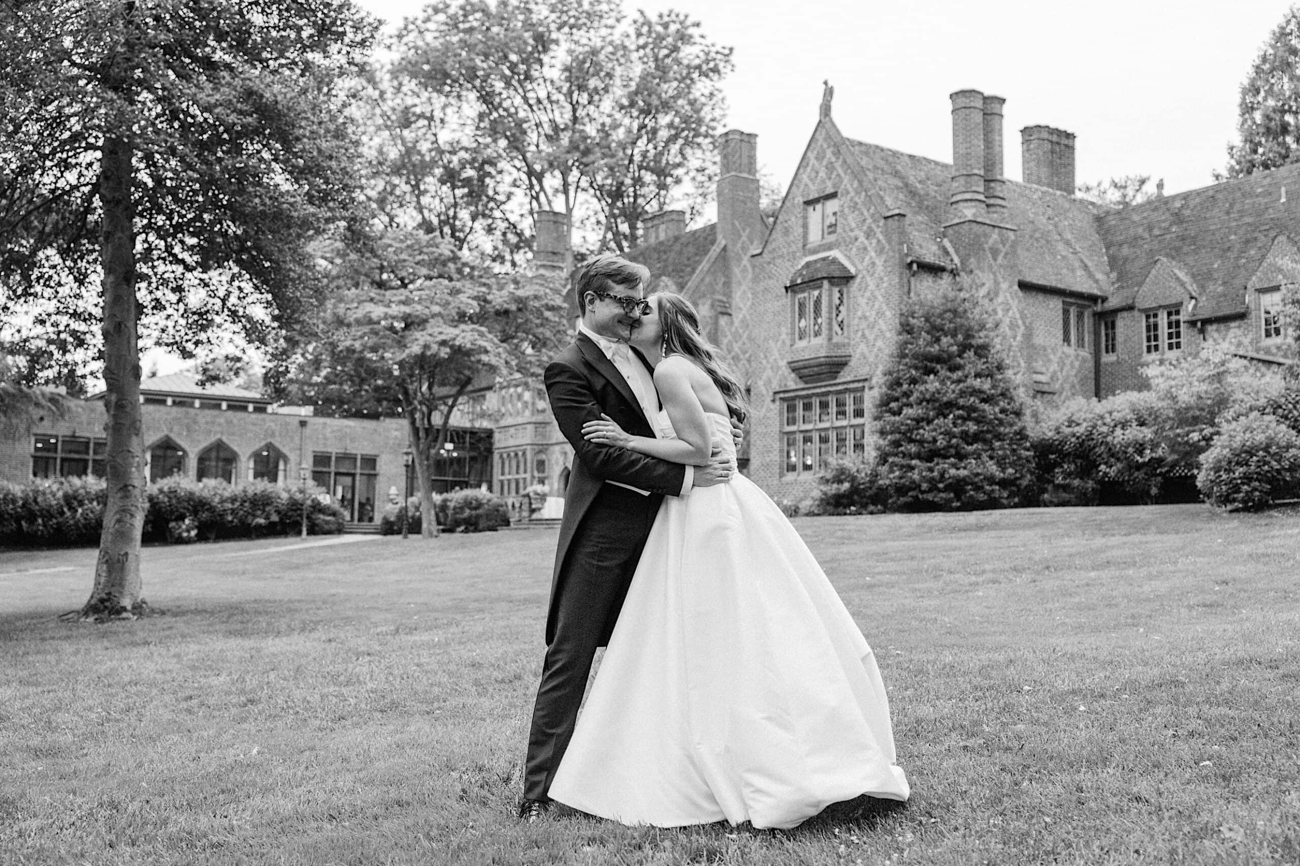 A newlywed couple kisses and embraces on a lawn in front of the historic Aldie Mansion, surrounded by trees—a picturesque moment from their wedding at Aldie Mansion.