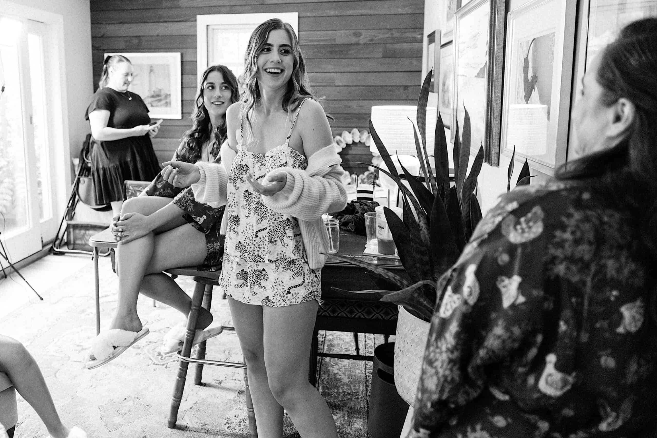 A group of women in casual attire chat and laugh in a cozy, well-lit room with framed photos on the wall and a large plant nearby, sharing memories of a recent wedding at Aldie Mansion.