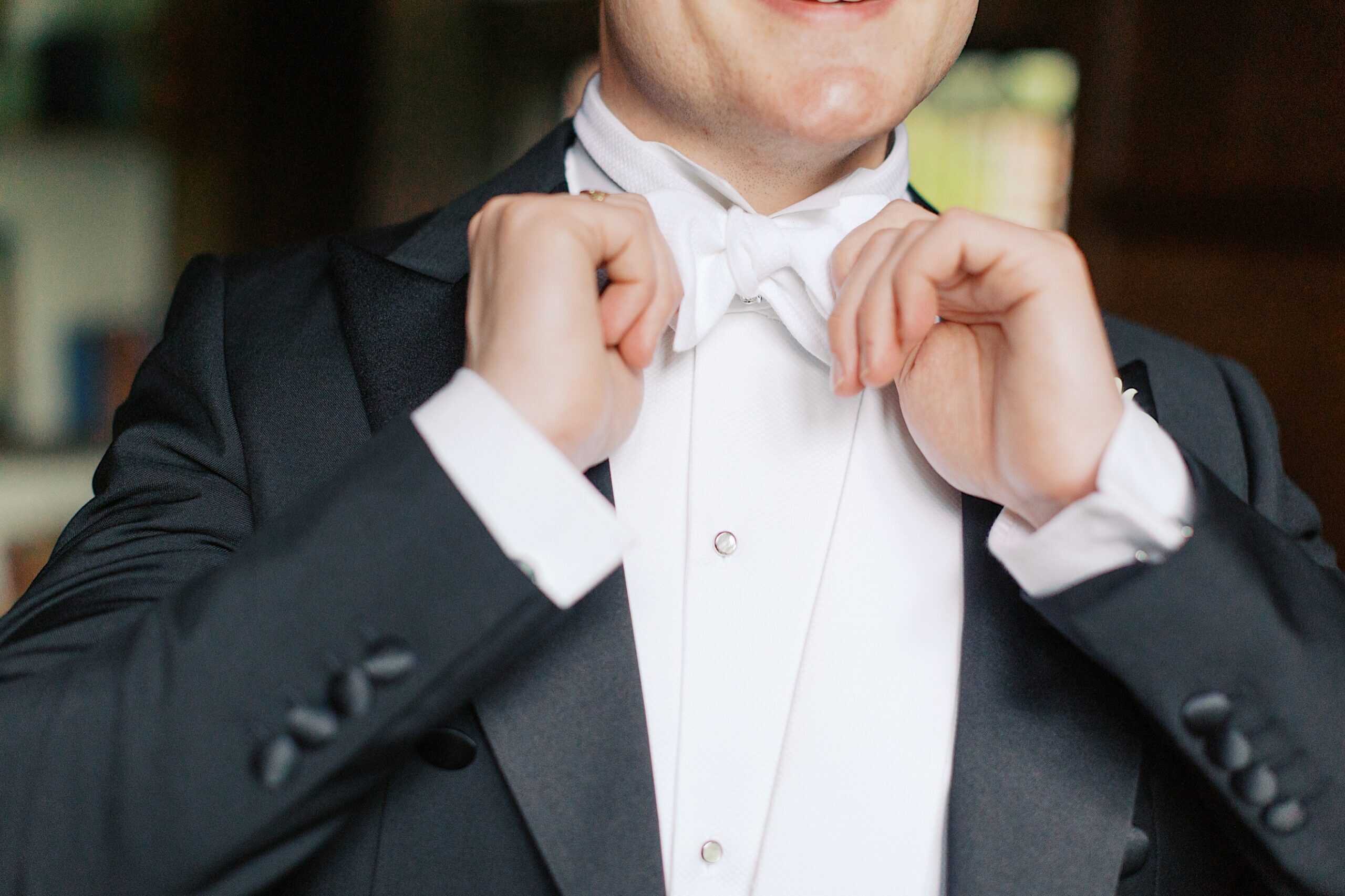 A person in formal attire adjusts a white bow tie while wearing a black tuxedo jacket and white dress shirt, preparing for a wedding at Aldie Mansion.