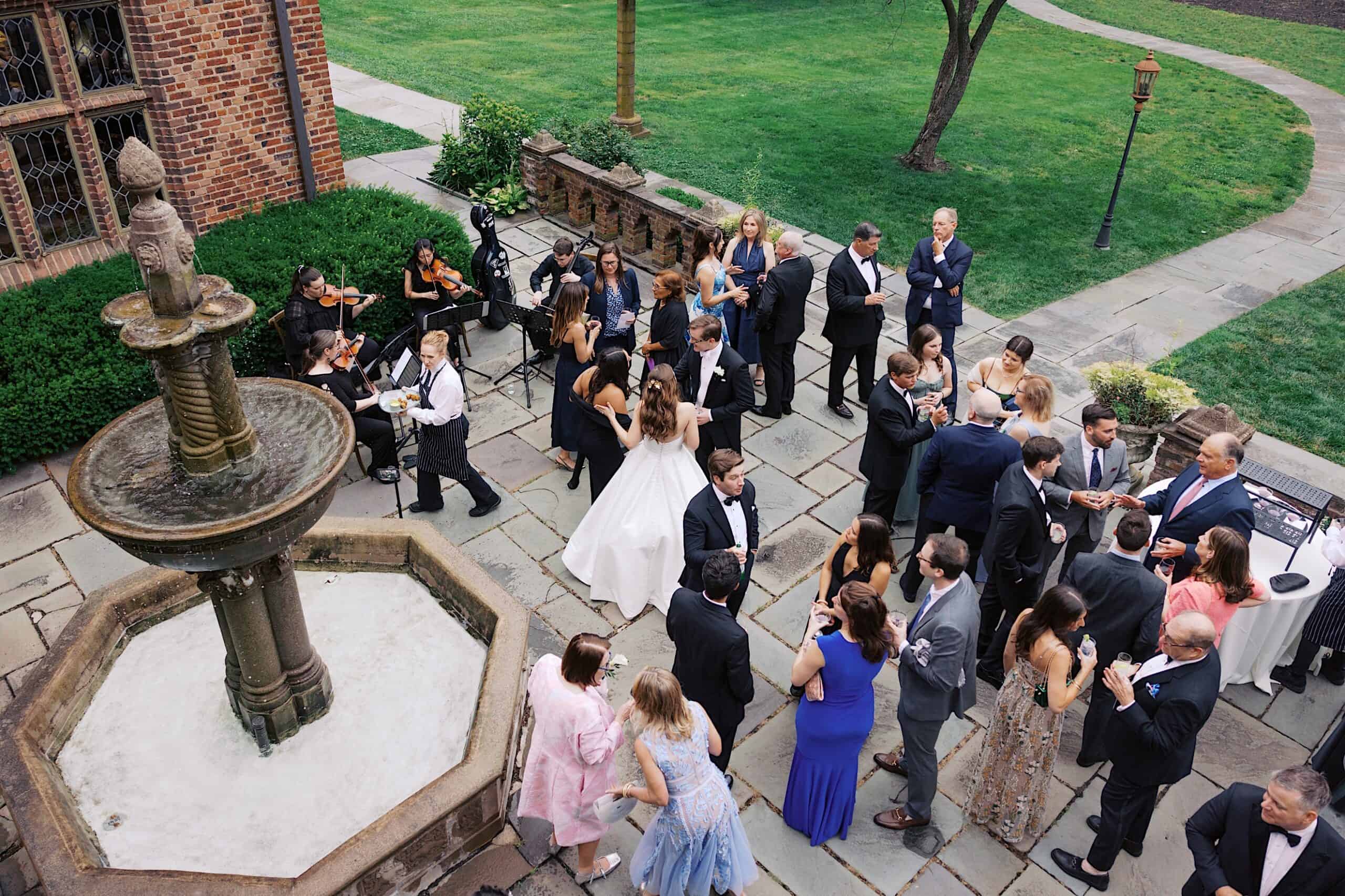 A group of people in formal attire gathers outdoors near a fountain for a social event, with a string quartet performing nearby at a beautiful Wedding at Aldie Mansion.
