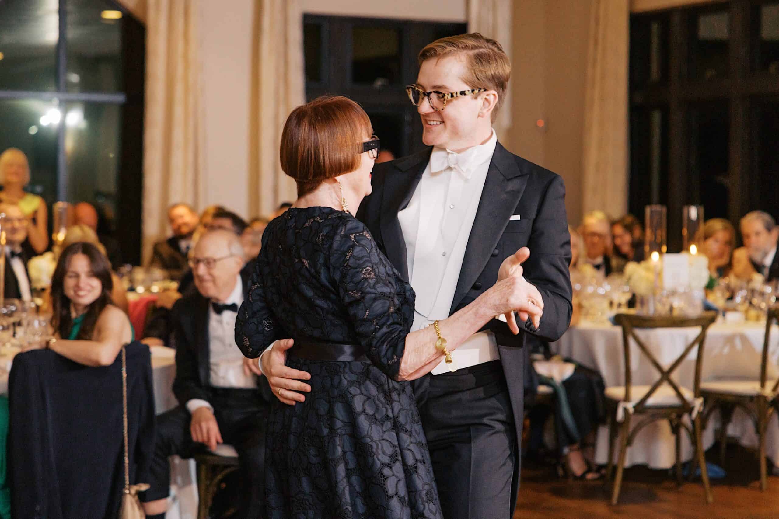 A man in formal attire and an older woman in a dark dress are dancing together at a Wedding at Aldie Mansion, with seated guests watching in the background.