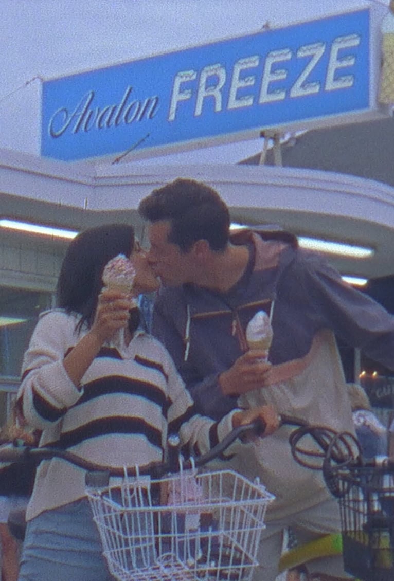 Two people stand with bicycles and ice cream cones, sharing a kiss in front of an ice cream shop with a sign that reads "Avalon Freeze.