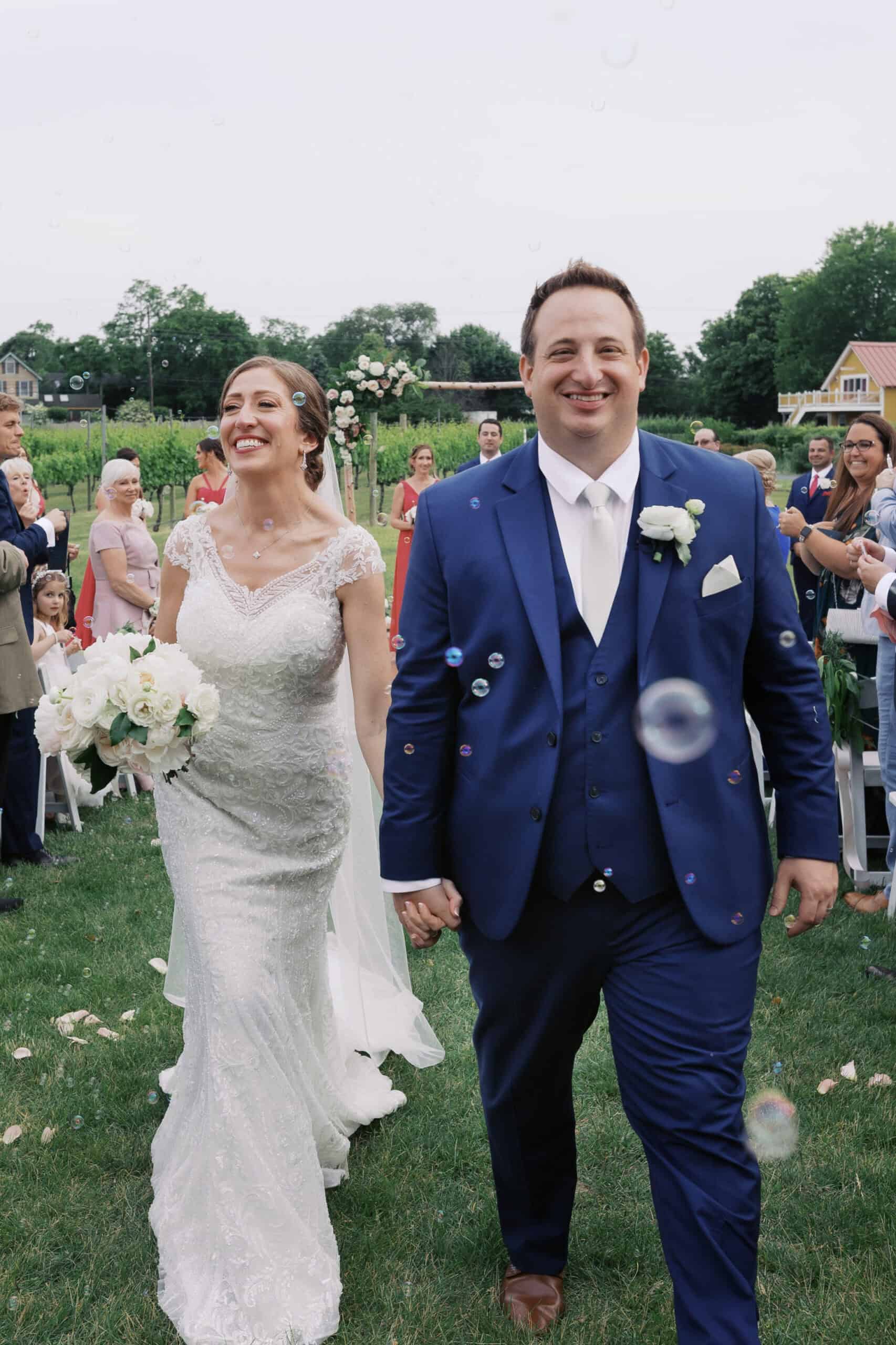 A bride in a white dress and a groom in a blue suit walk outdoors, smiling and holding hands, surrounded by guests and bubbles at a wedding ceremony.
