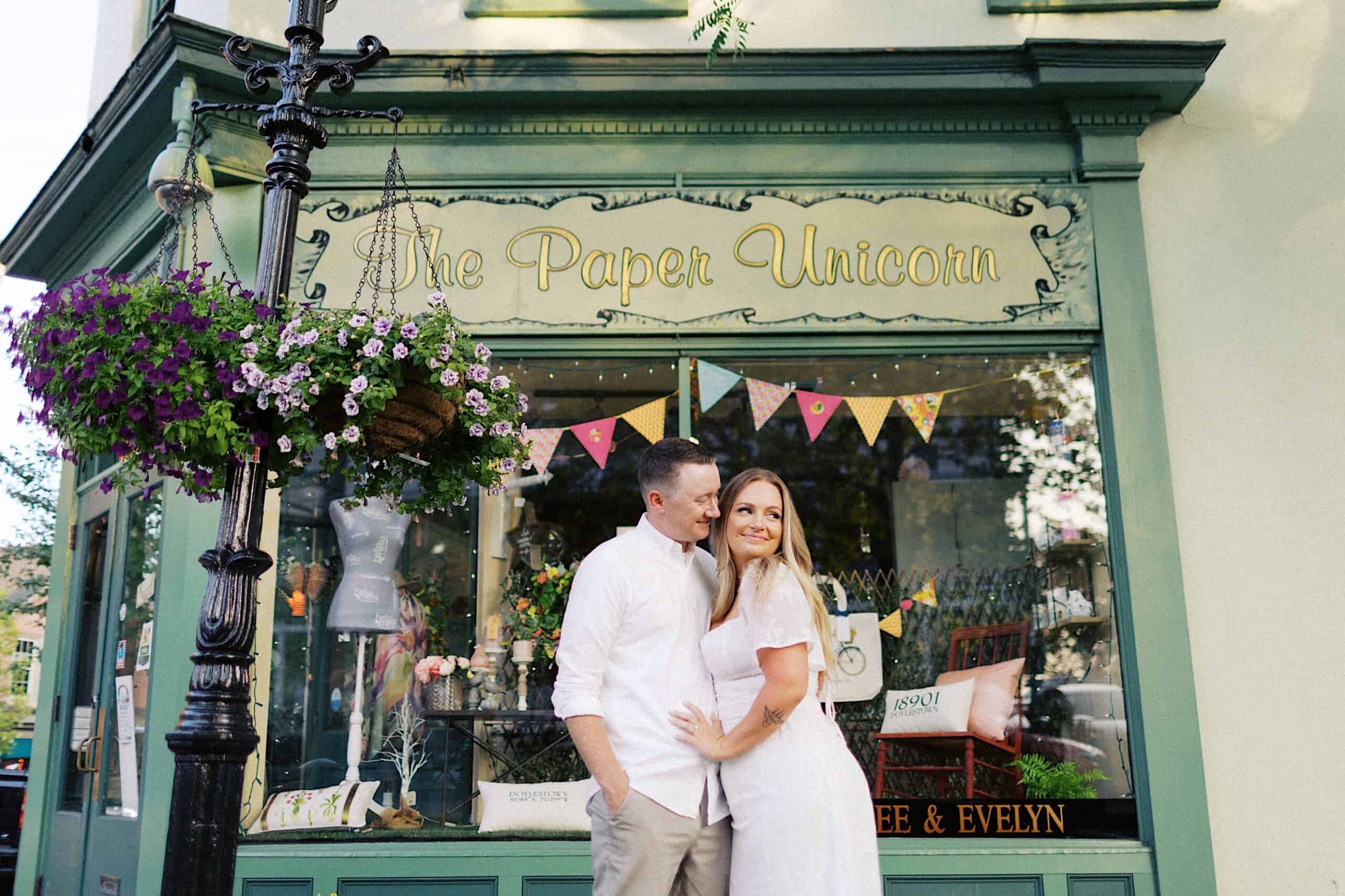 A couple in white outfits stands smiling closely in front of a shop called "The Paper Unicorn," with decorative flowers and bunting visible in the window.