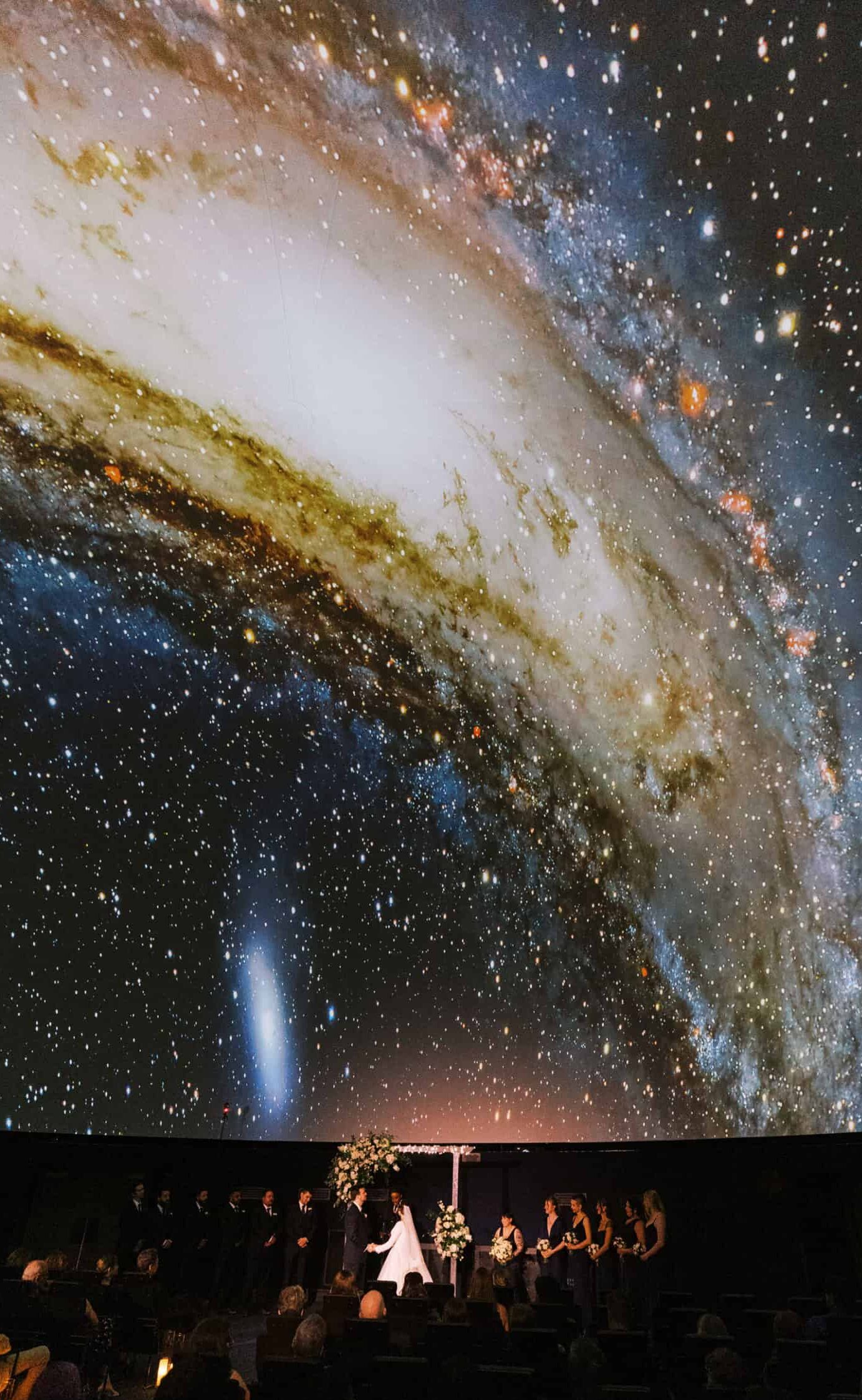 A wedding ceremony takes place in a theater, with a large galaxy projected on the domed ceiling above the couple and guests—a magical setting reminiscent of a Franklin Institute wedding.