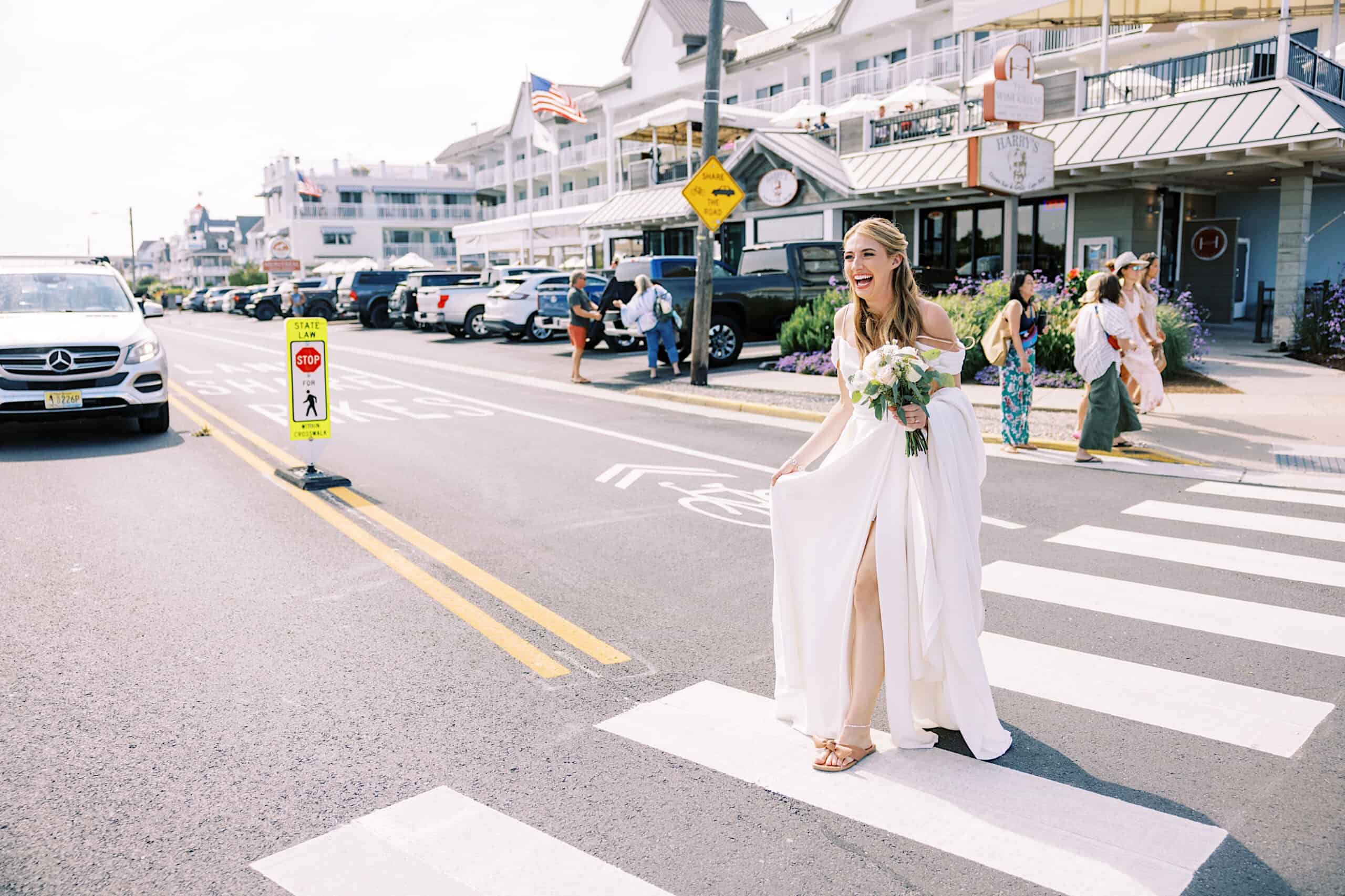 A woman in a white dress holding a bouquet walks across a crosswalk near parked cars and buildings, capturing the elegant vibe of a Montreal Beach Resort Wedding with people in the background.