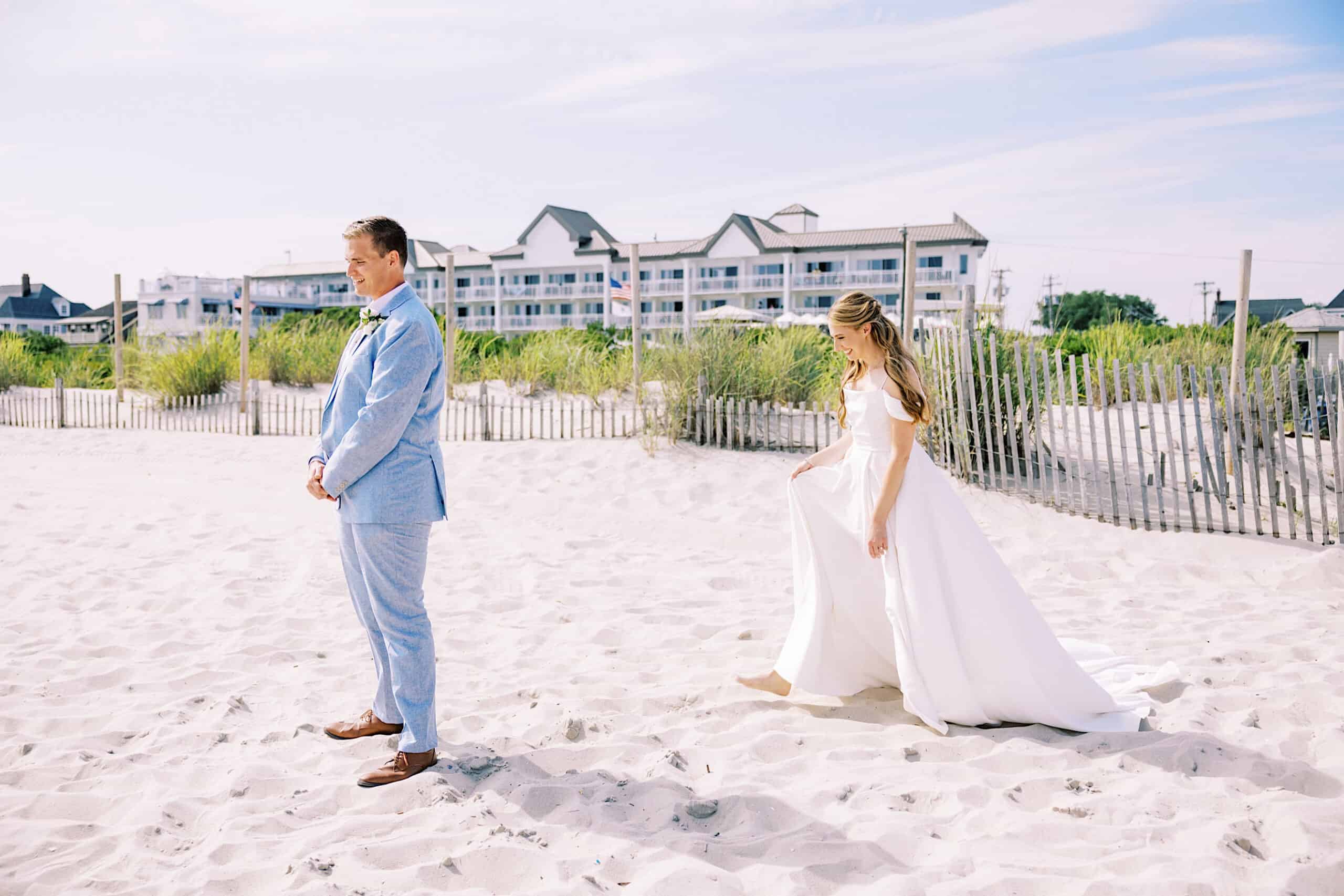 A bride in a white dress approaches a groom in a light blue suit on a sandy beach at the Montreal Beach Resort, with buildings and a wooden fence in the background.
