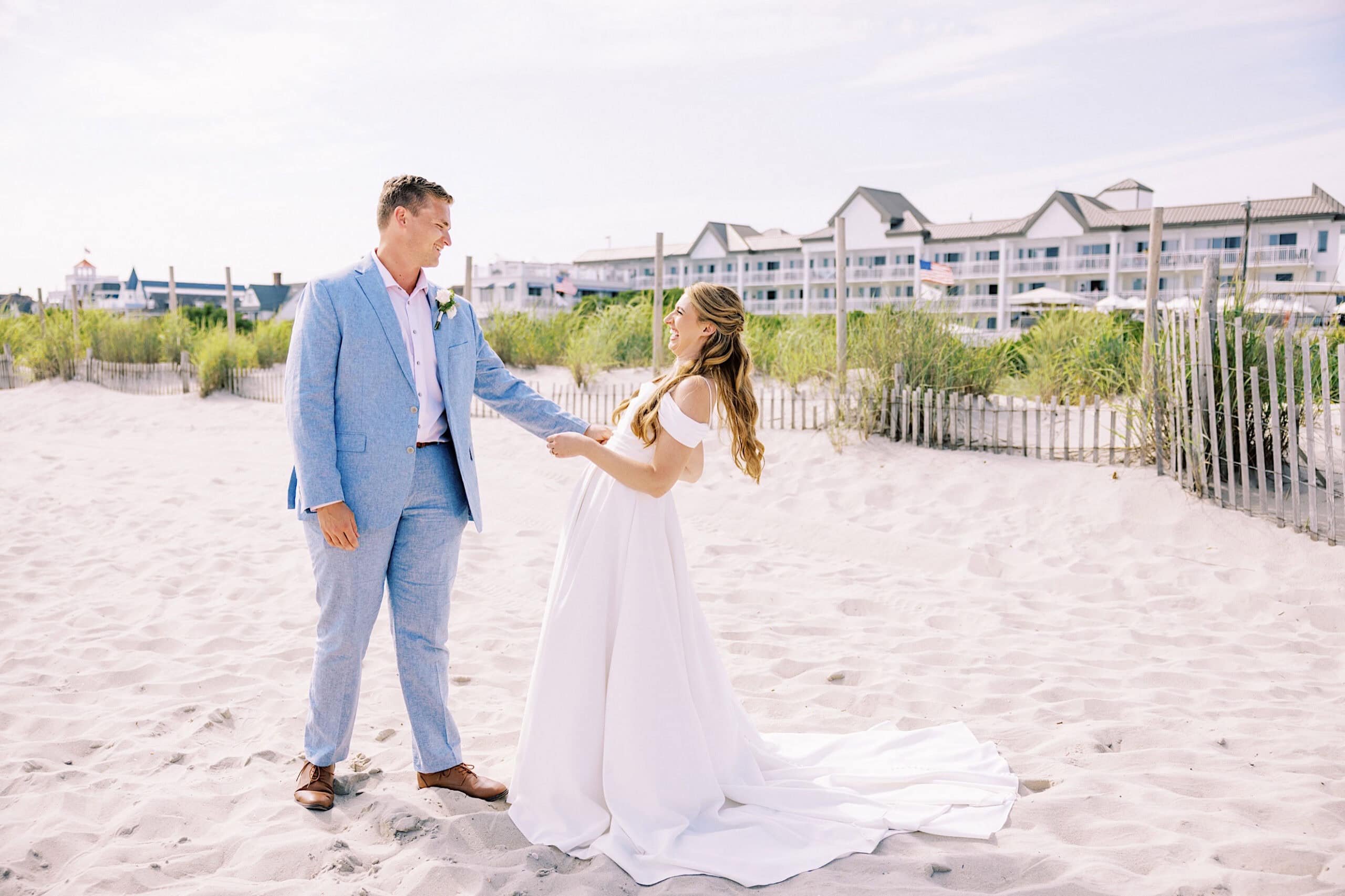 A bride and groom stand on a sandy beach at their Montreal Beach Resort wedding, holding hands and smiling at each other in front of beach grass and charming buildings.